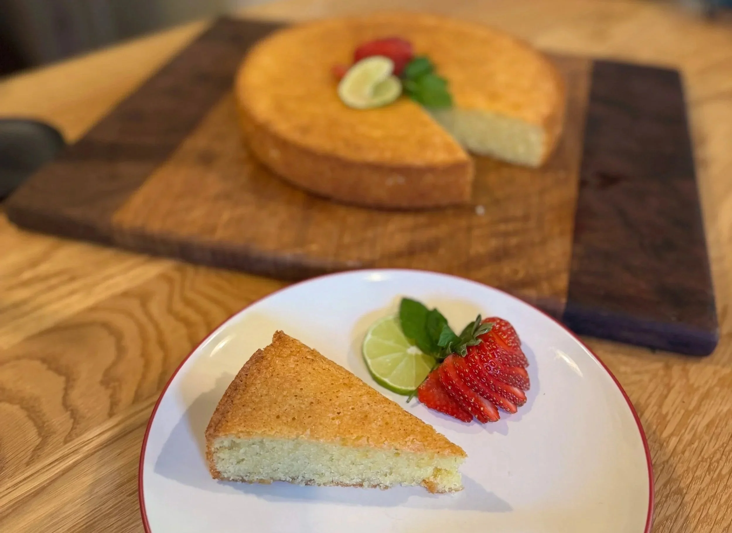 A slice of sponge cake on a white plate with sliced strawberries, a lime wedge, and a mint leaf. A whole layered cake with lime slices, strawberries, and mint leaves is on a wooden serving board in the background.