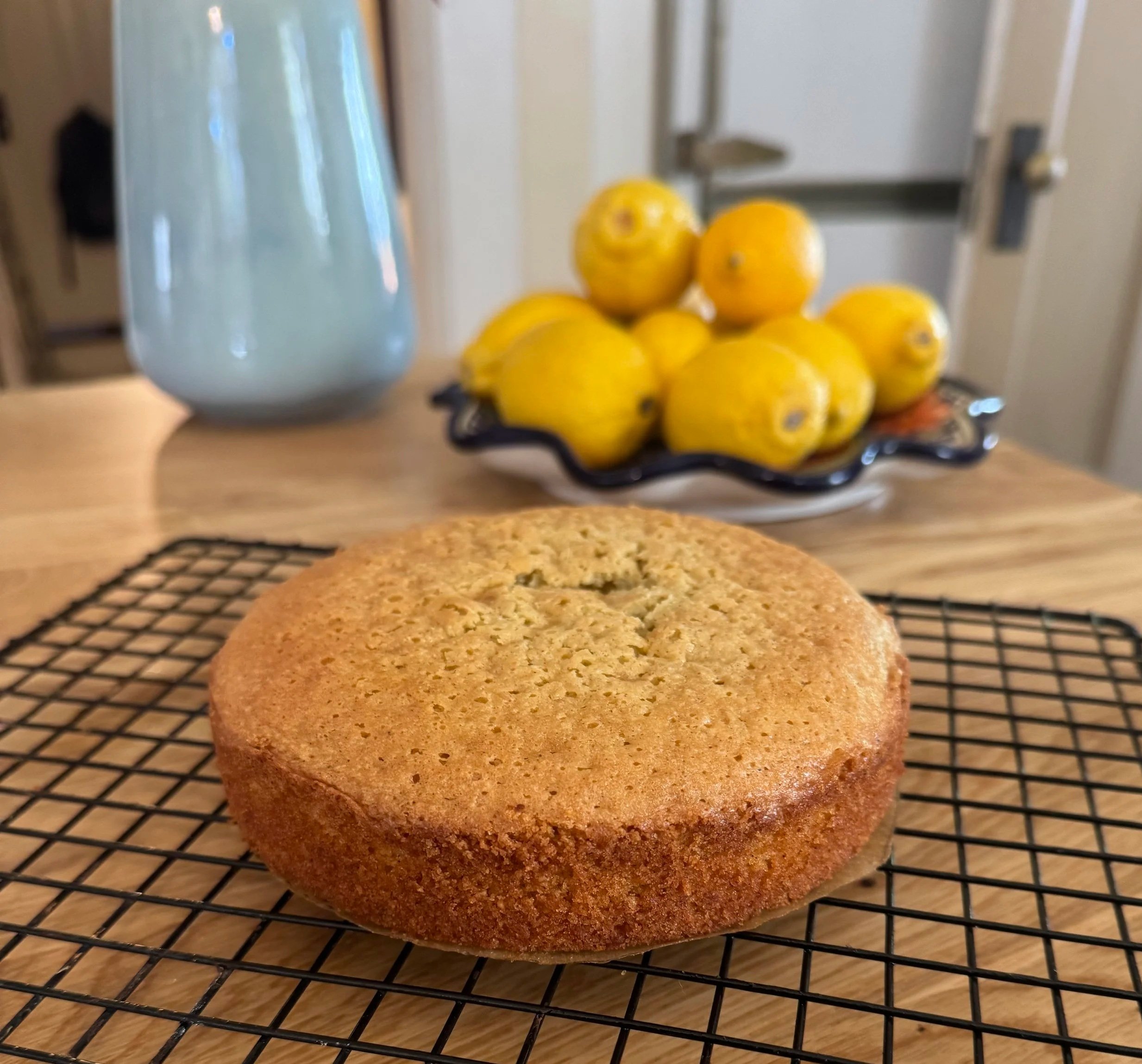 A freshly baked round cake cooling on a wire rack in the foreground, with a plate of yellow lemons and a large light blue vase in the background on a wooden table.