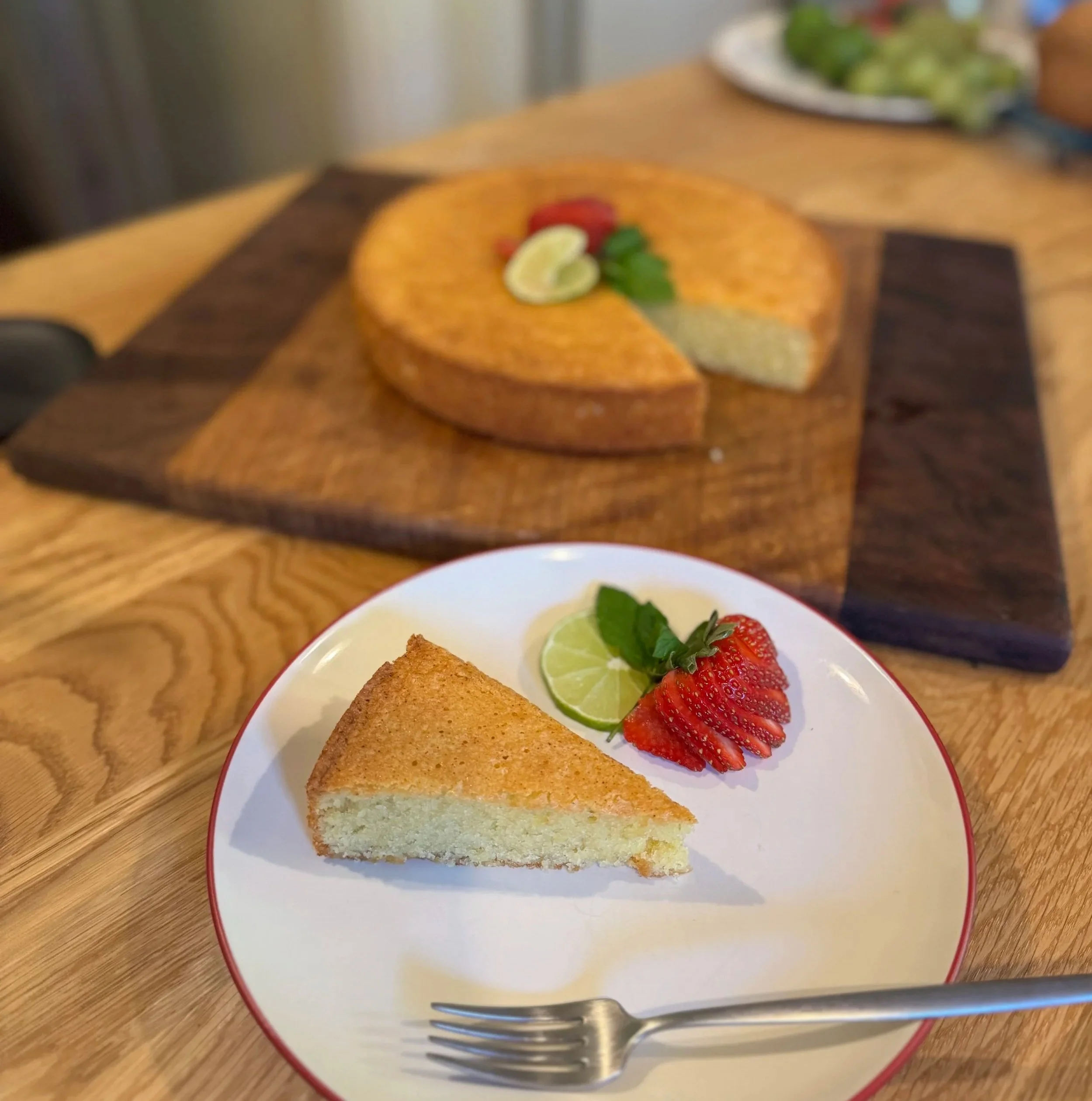 A slice of gluten free, dairy-free almond olive oil lemon cake with a golden brown crust, garnished with a lemon slice, a strawberry, and mint leaves, on a white plate with a fork in front.
