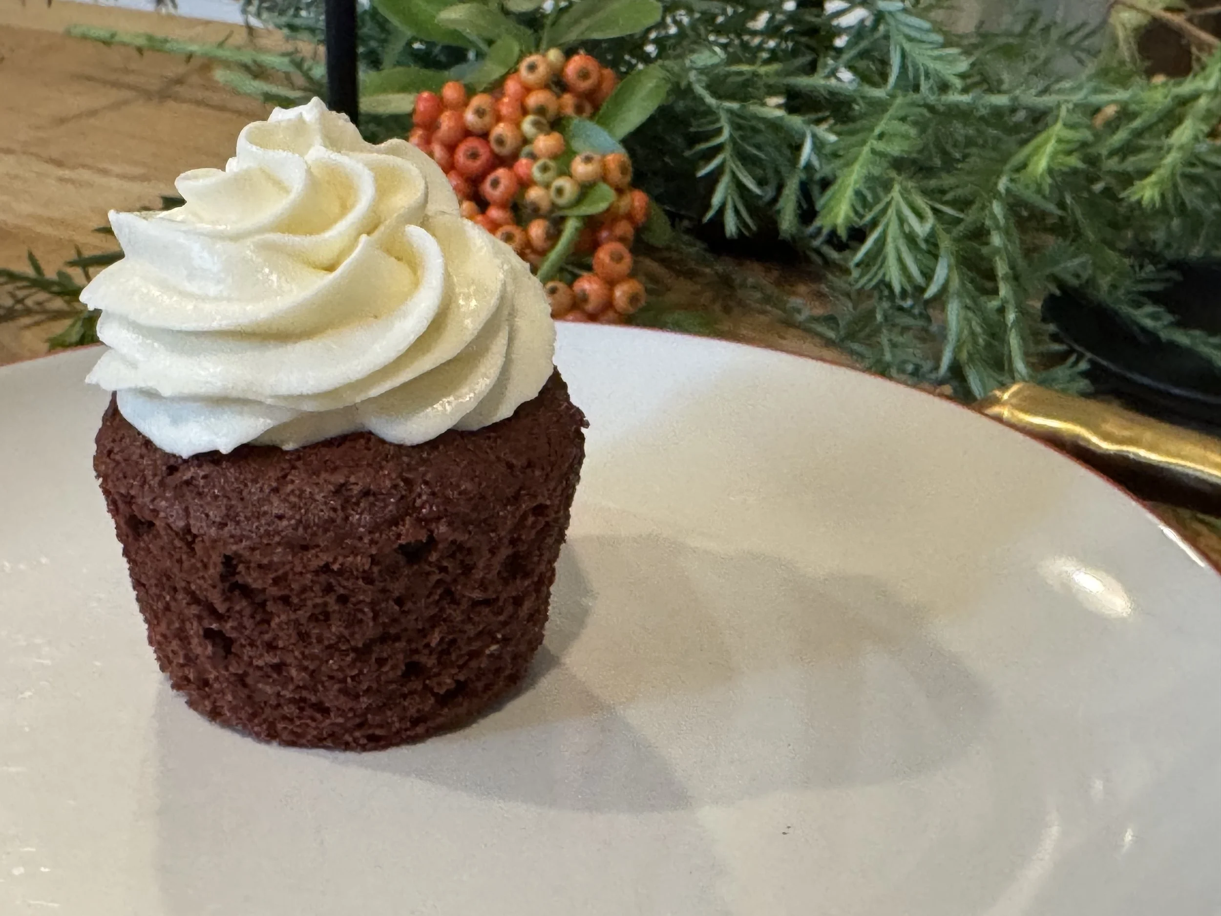 A chocolate cupcake with white frosting swirl on top, placed on a white plate. In the background, there are sprigs of greenery and small orange berries, suggesting a festive or holiday setting.