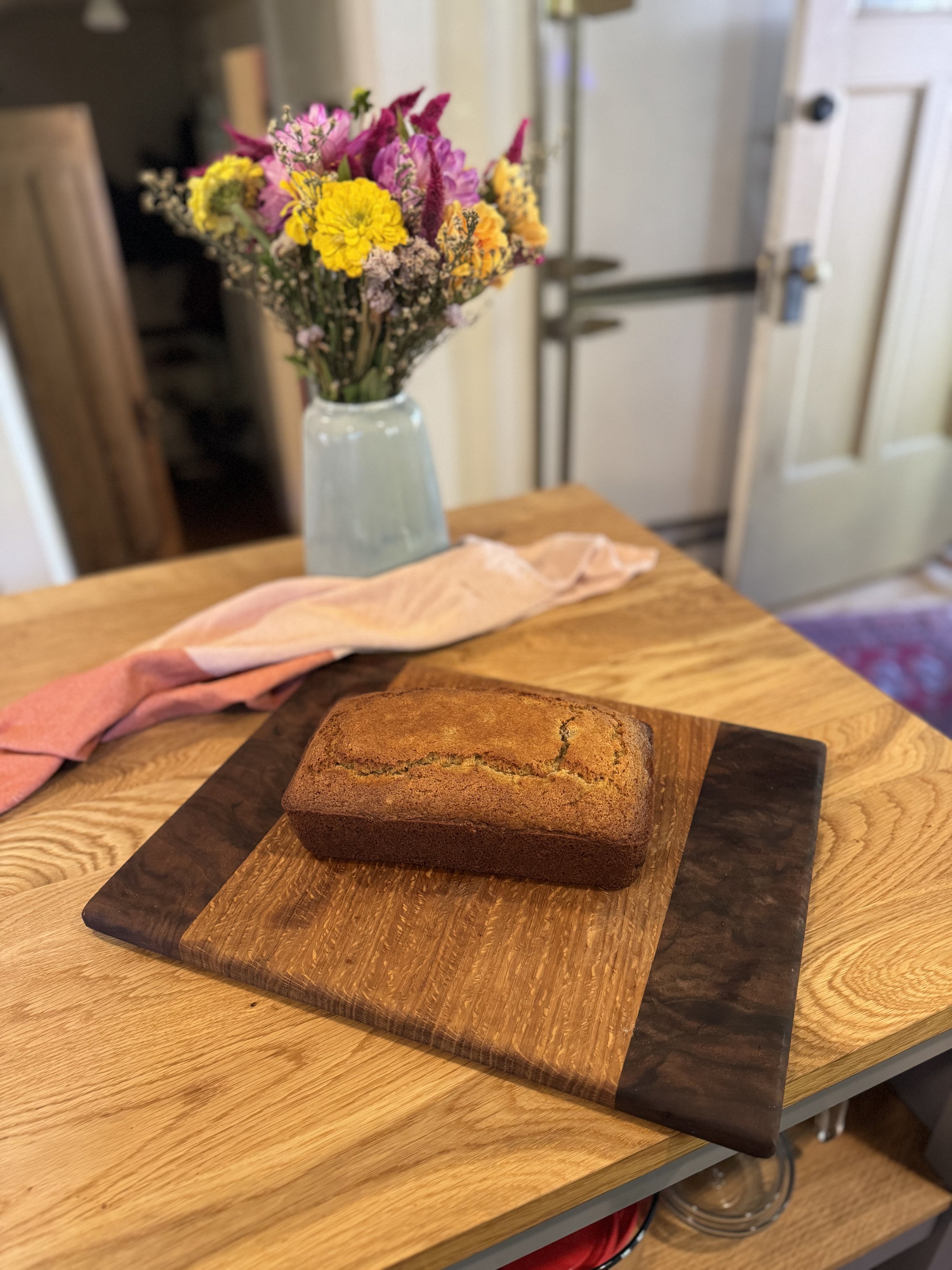 Freshly baked loaf of banana bread on a wooden cutting board on a kitchen table with a pink and orange cloth and a vase of multicolored flowers in the background.