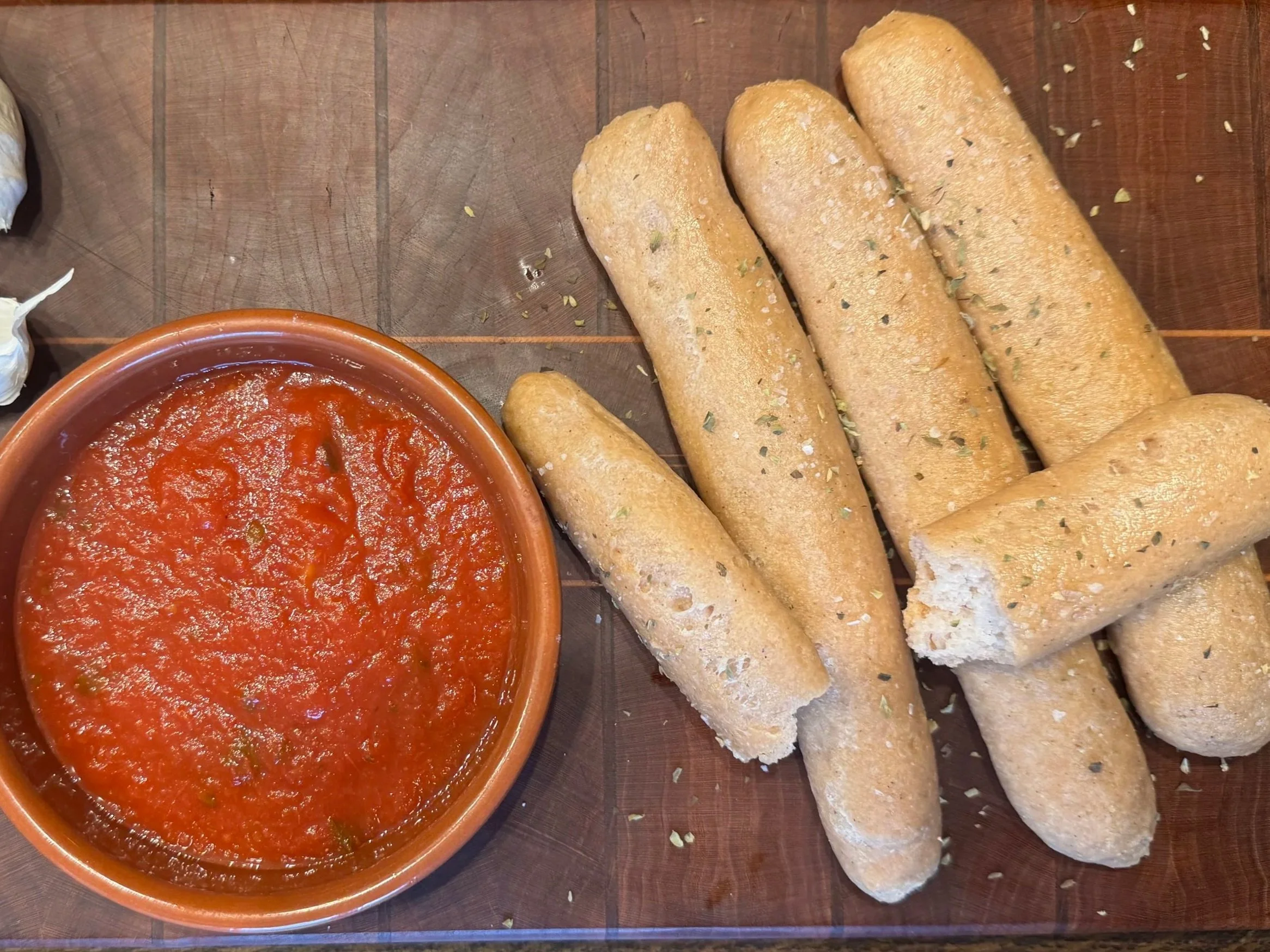 A bowl of red tomato sauce and several pieces of toasted bread sticks on a wooden serving board.