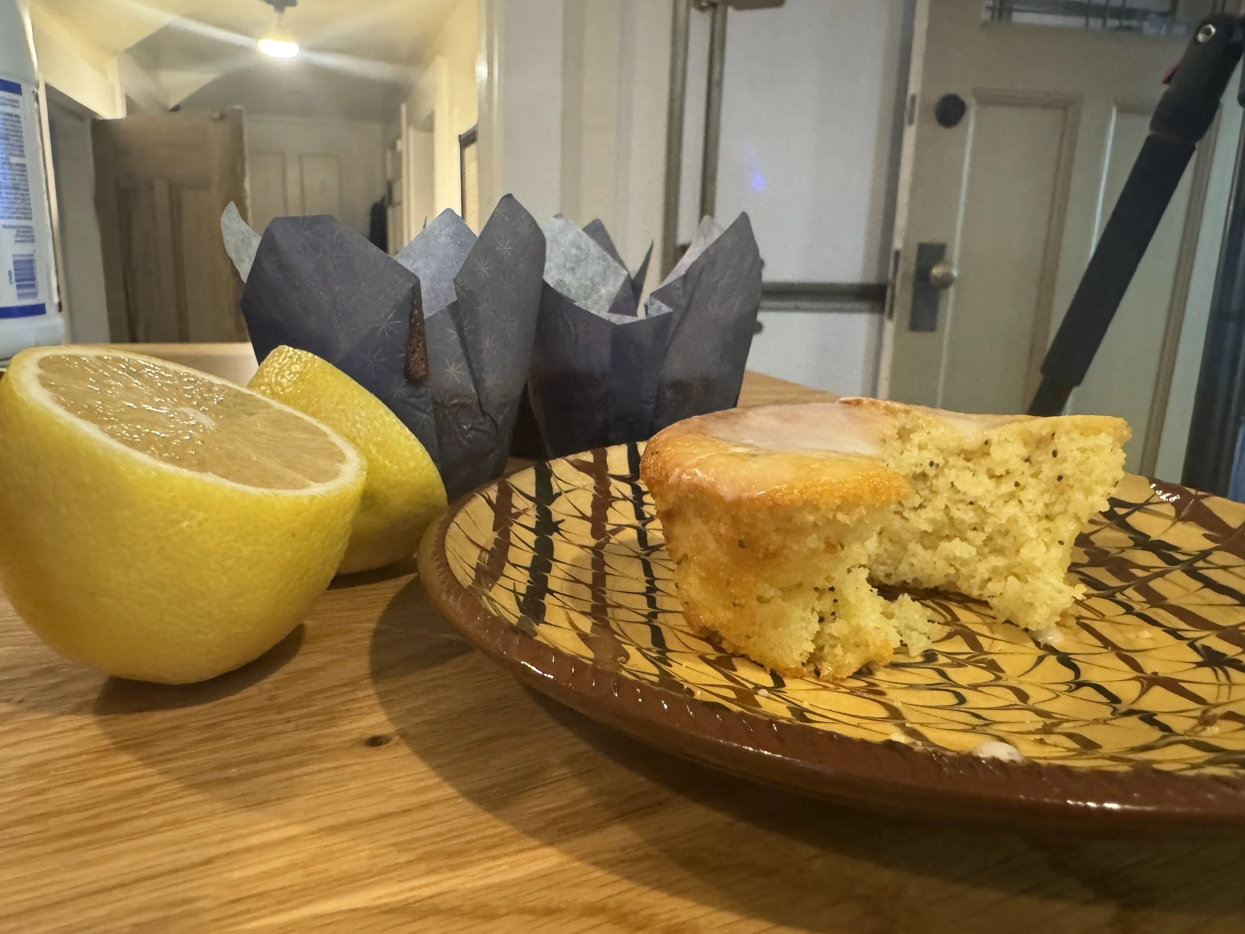 A partially eaten slice of lemon cake on a decorative plate, with two lemon halves and a paper cupcake wrapper behind it on a wooden table.