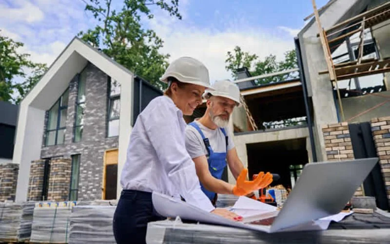 Two construction workers, a woman and a man, wearing safety helmets inspect plans at a construction site for a modern house.