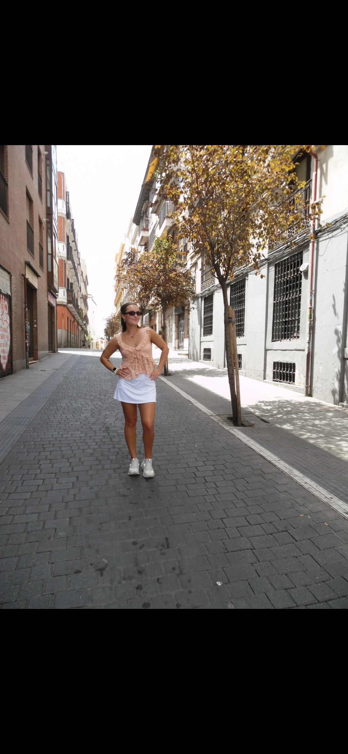 A woman standing on a cobblestone street lined with trees and urban buildings, wearing sunglasses, a peach top, a white skirt, and athletic shoes.