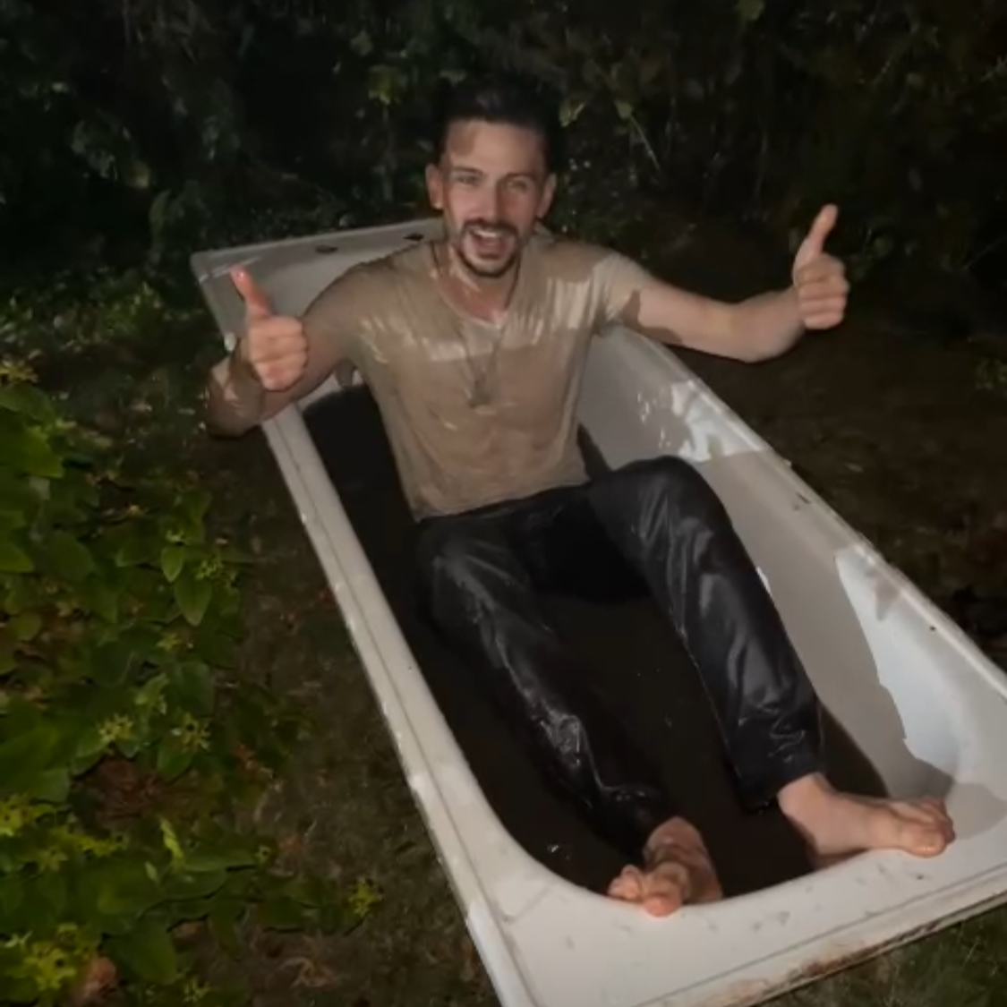 A man sitting in an outdoor bathtub, covered in mud, giving a thumbs-up with both hands, at night surrounded by greenery.