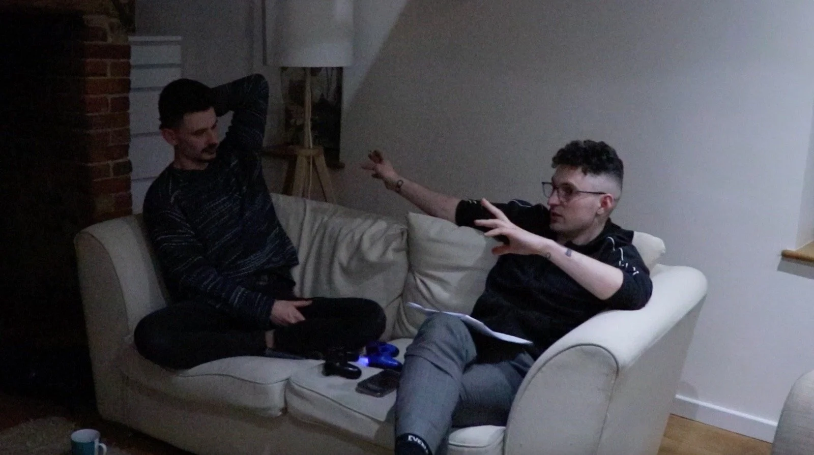 Two young men sit on a cream-colored sofa in a dimly lit room, one with a beard and dark hair, the other with glasses and curly hair, engaging in a conversation.