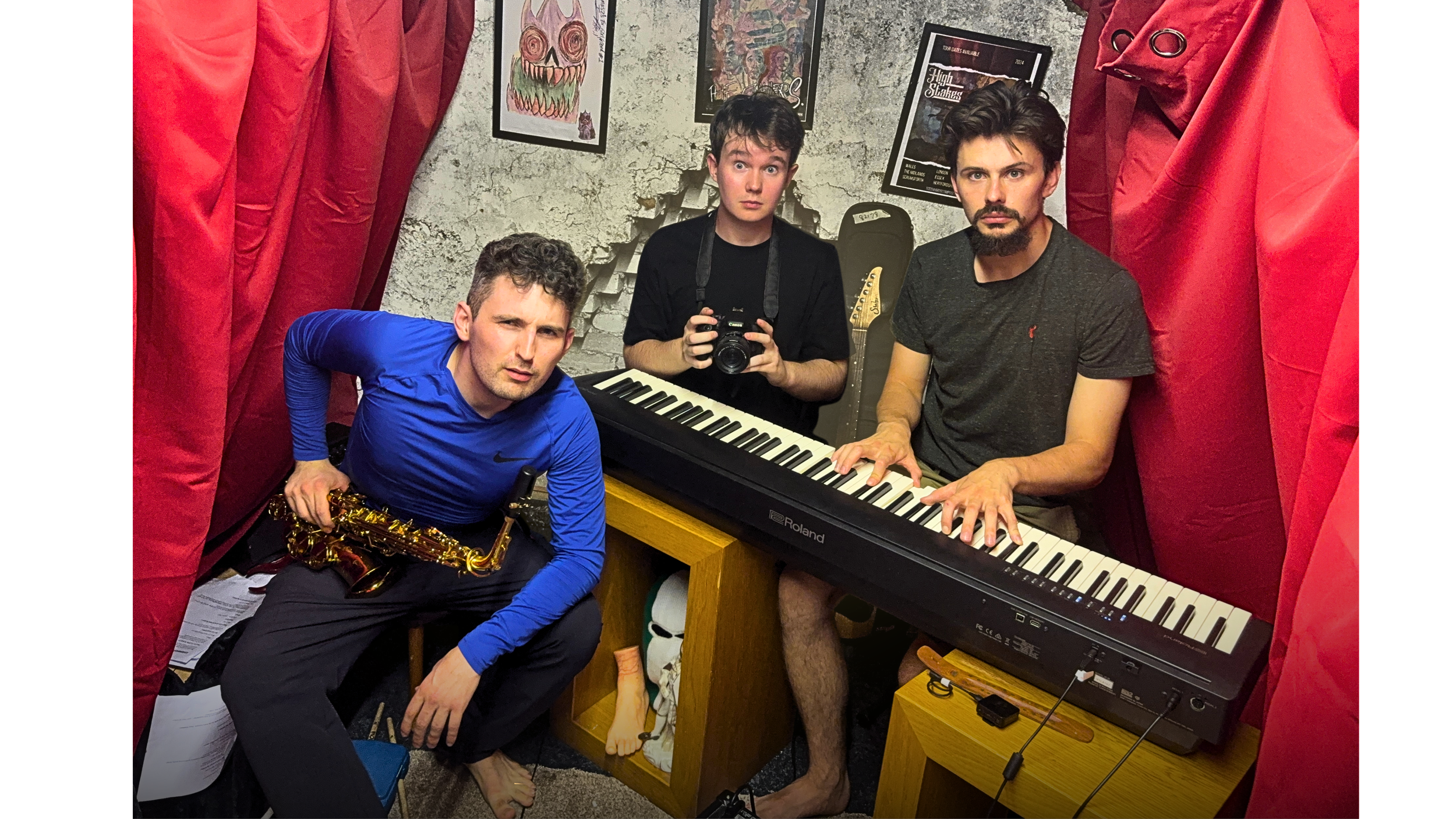 Three men in a small music room with red curtains, posters on the wall, and a keyboard. One man is playing the keyboard, another is holding a saxophone, and the third man is taking a photo. The man on the right has a beard and is wearing a dark t-shirt, the man in the middle is holding a camera, and the man on the left has curly hair and is dressed in blue.