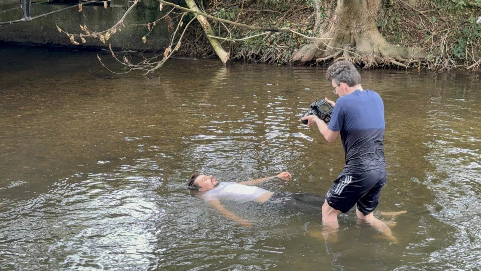 A man is floating in a shallow body of water while another man takes photos of him. The background shows trees and roots along the shore.