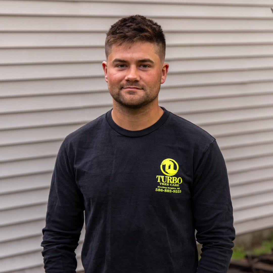 A young man with short brown hair and a beard, wearing a black long-sleeve shirt with a yellow logo and text that reads "Turbo Tree Care" and a phone number. He is standing outdoors in front of gray siding.