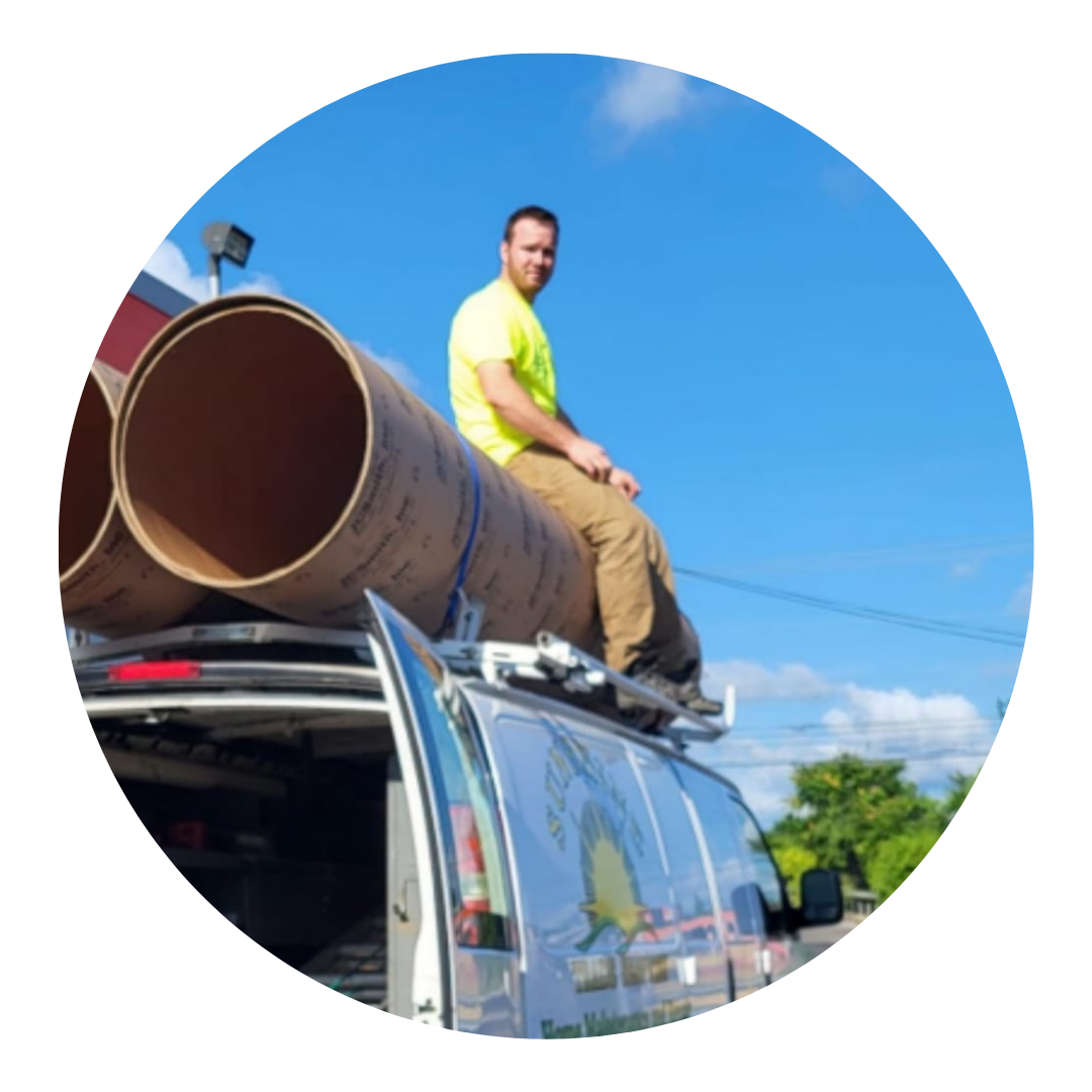 A man wearing a yellow shirt sitting on top of a van with large pipes loaded on the roof, under a blue sky with some clouds and trees in the background.