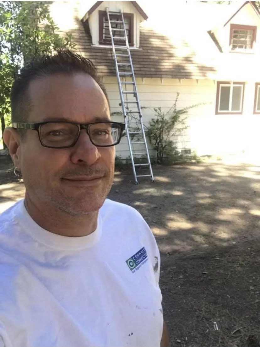 Man taking a selfie outdoors in front of a house with a ladder leaning against the upper window.