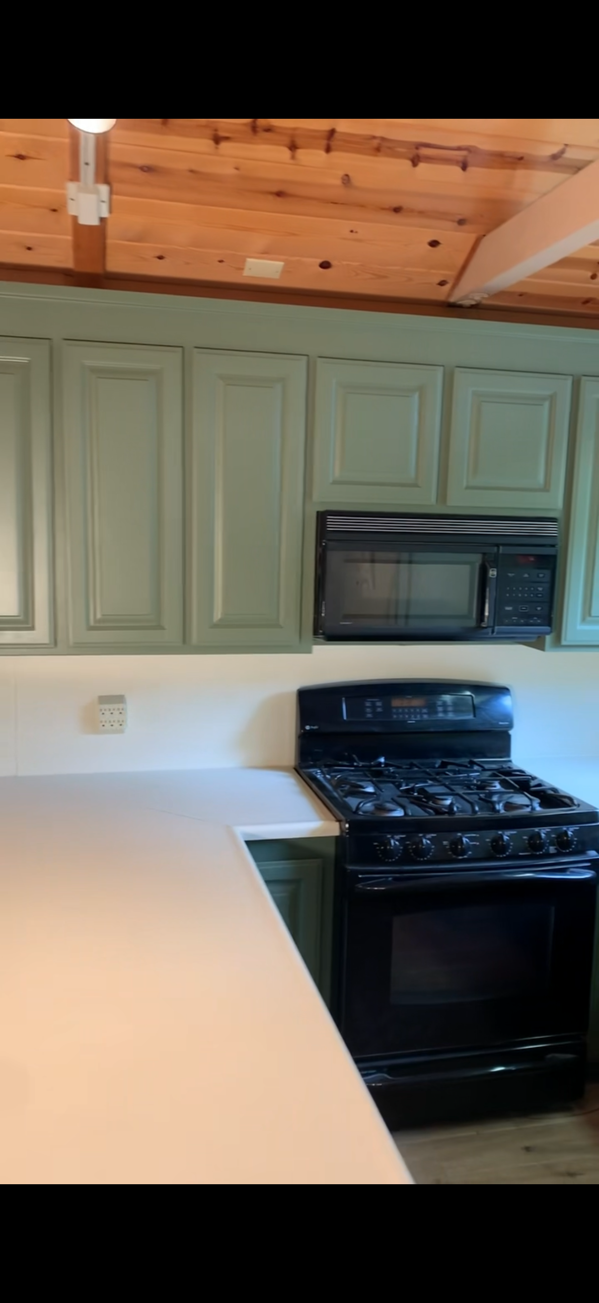 Kitchen with green cabinets, black microwave above a black stove, white countertops, wooden ceiling, and a small wall-mounted device.