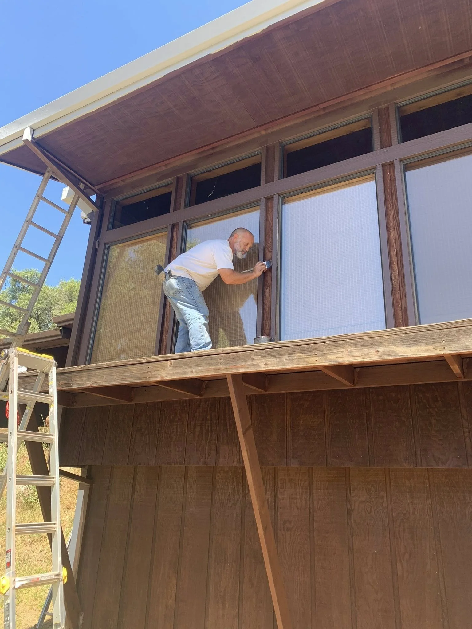A man standing on a wooden balcony, painting or working on the window frame of a modern house, with a ladder nearby and trees in the background.