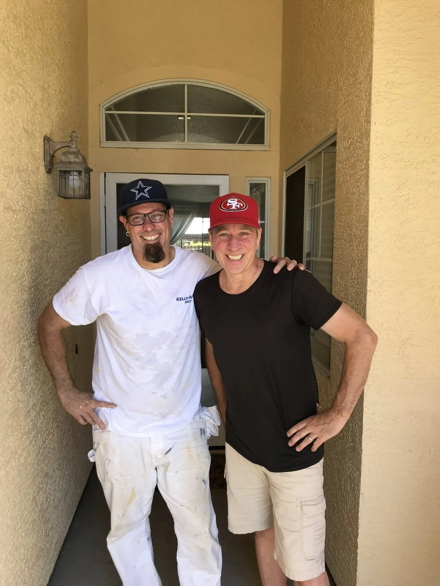 Two smiling men standing on a porch with yellow walls, wearing casual clothes and baseball caps, one with a Dallas Cowboys cap and the other with a 49ers cap.