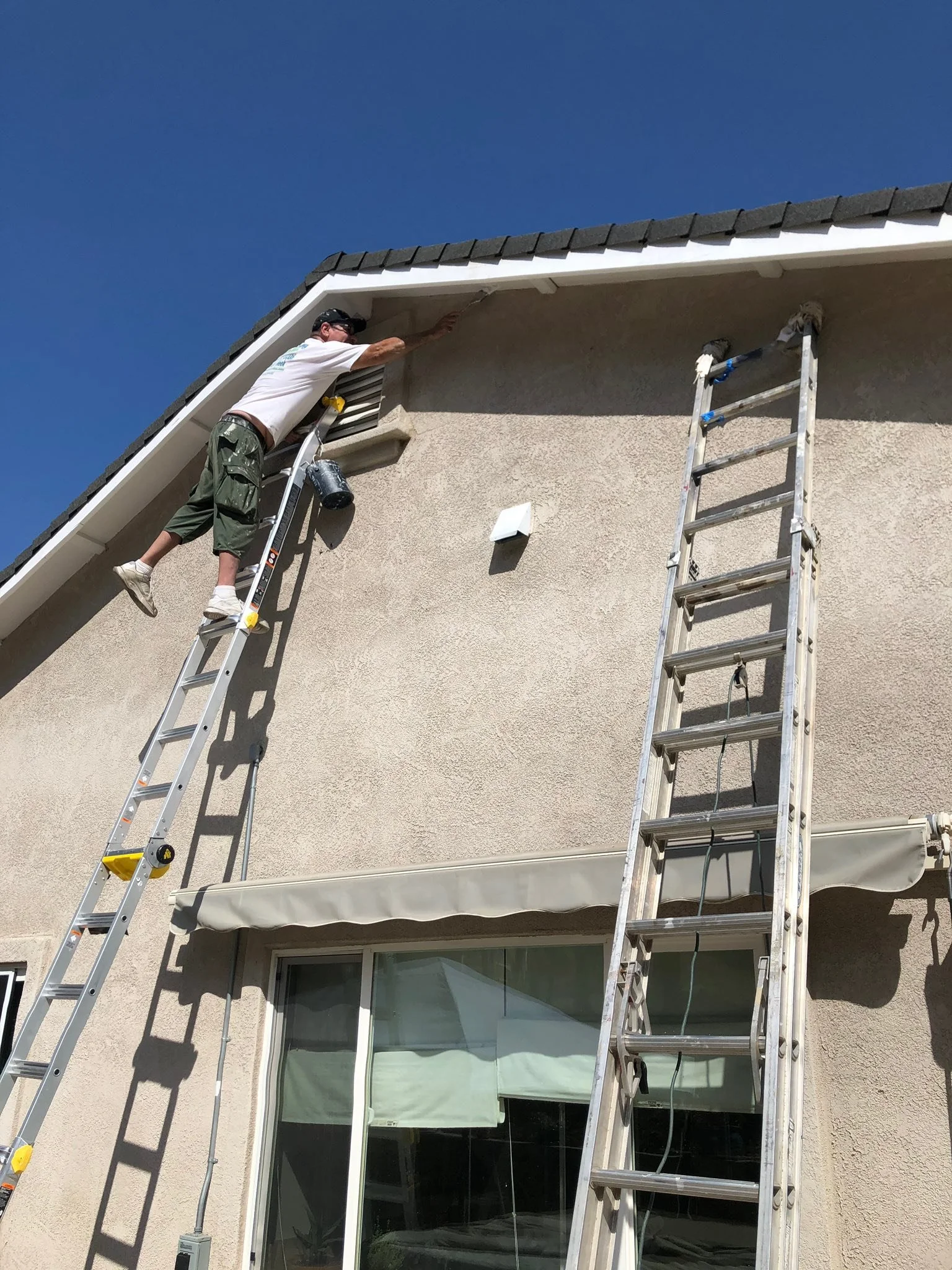 A man standing on a ladder cleaning or inspecting the exterior roof of a house under a clear blue sky, with a second ladder leaning against the house.
