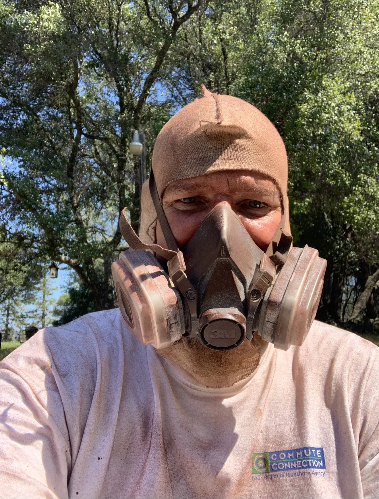Man wearing a tan work cap and a protective 3M mask, outdoors with trees and blue sky in the background.