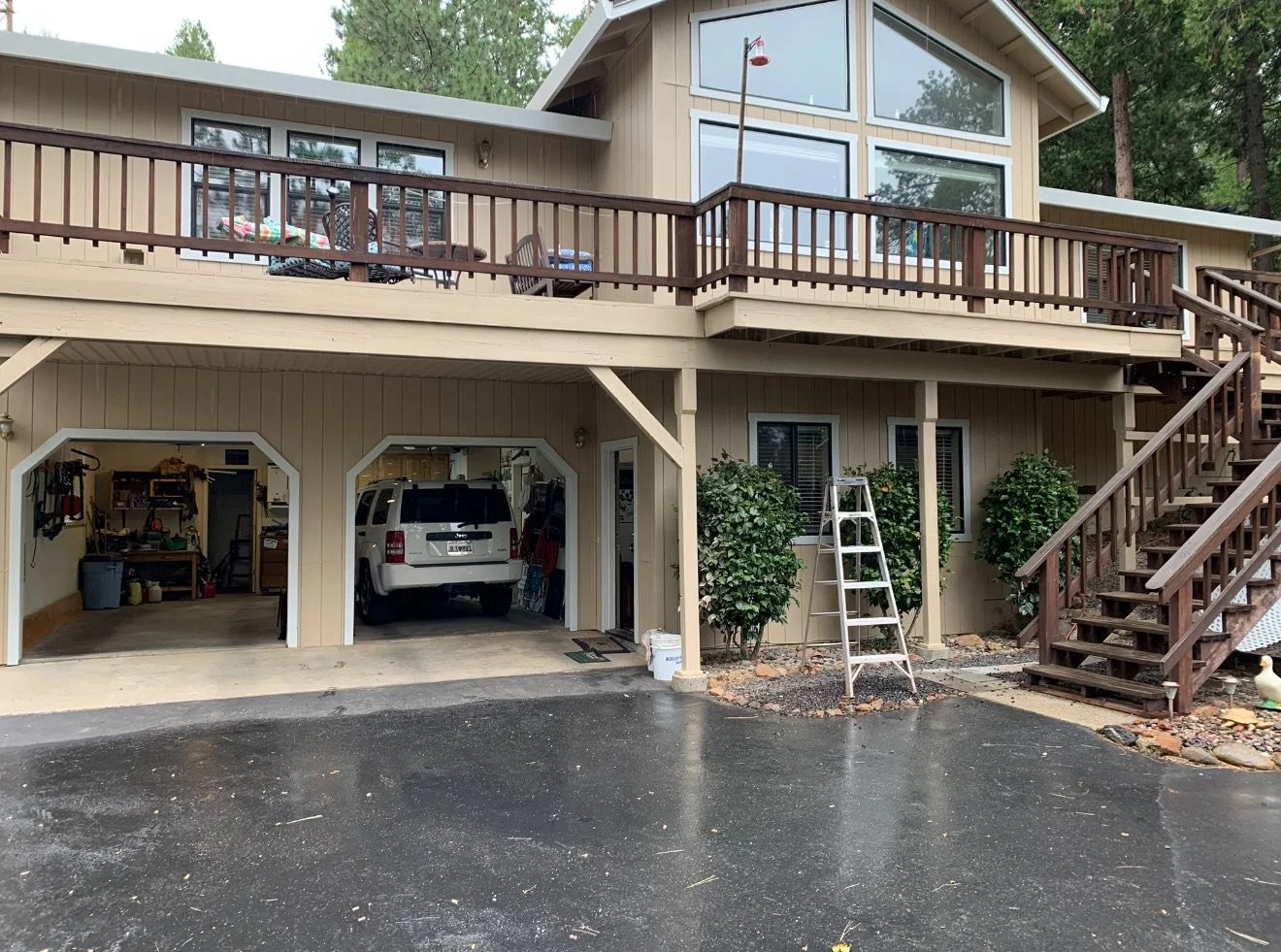 Two-car garage with one vehicle parked inside and an open door, beige house with large upper windows, wooden deck with outdoor furniture, ladder, stairs, and surrounding greenery.