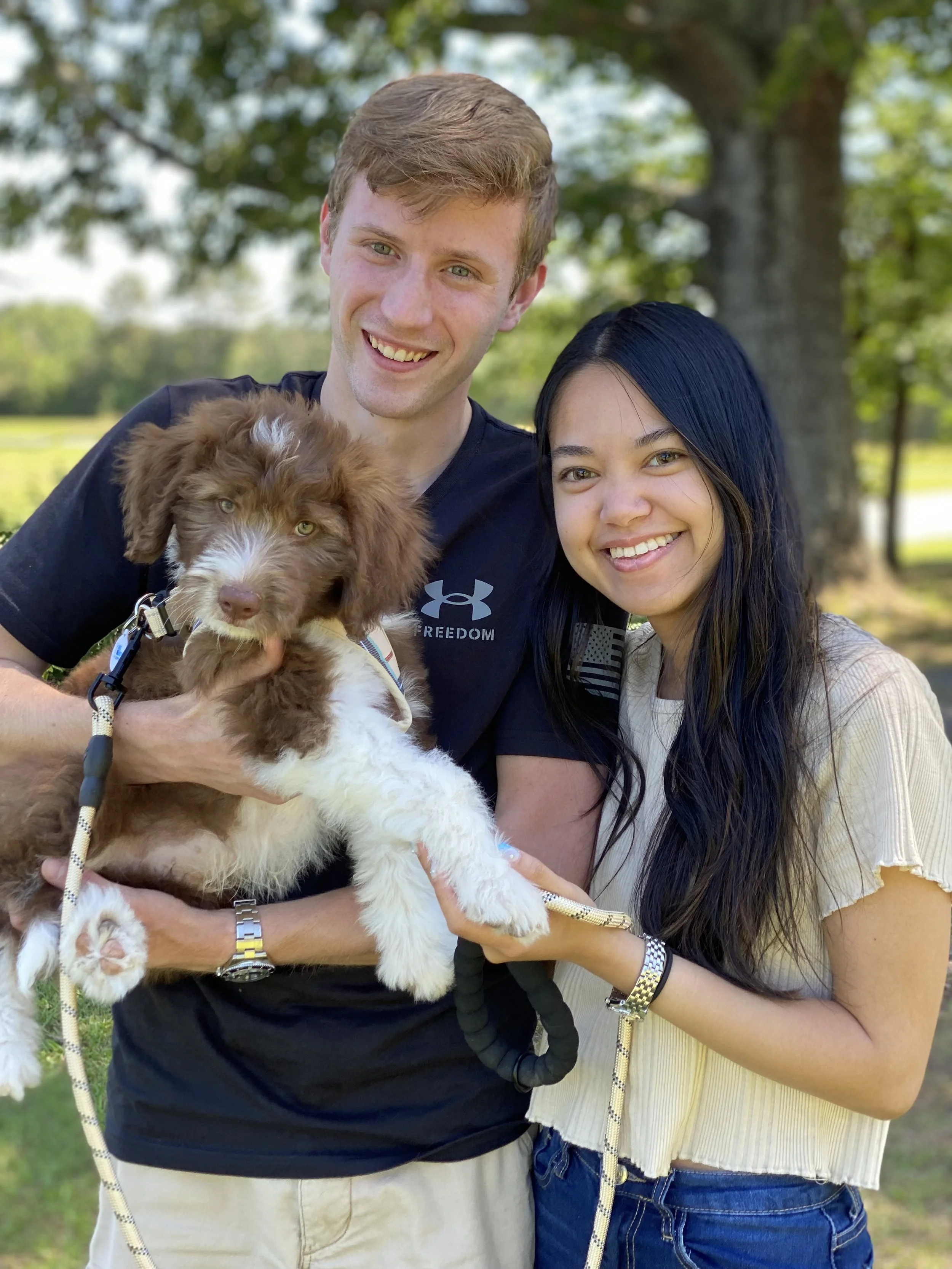 A smiling young man with light brown hair and a young woman with long dark hair, both holding a brown and white puppy outdoors with trees in the background.