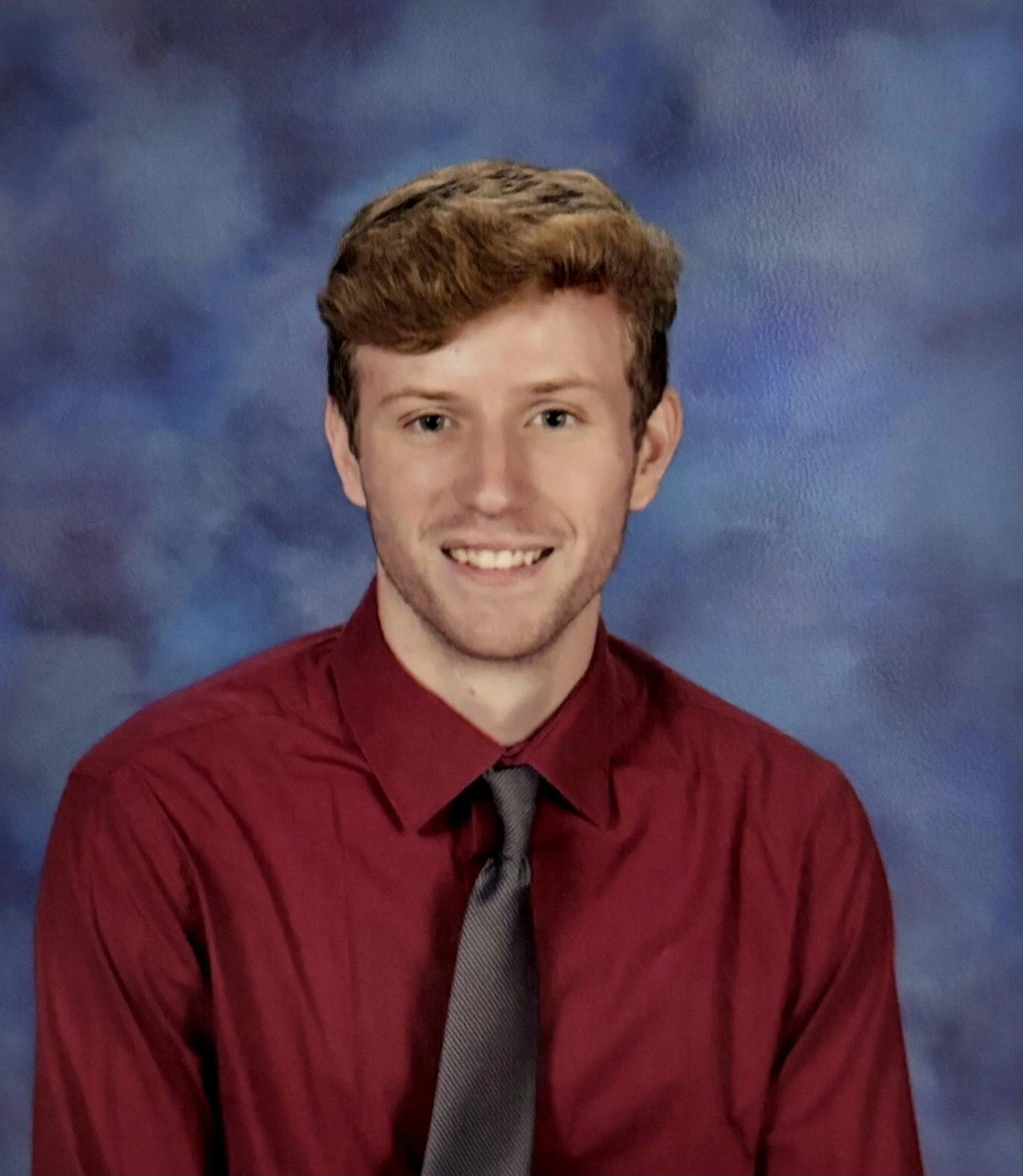 A young man with light brown hair, wearing a red dress shirt and a dark tie, smiling in front of a blue and purple cloudy background.