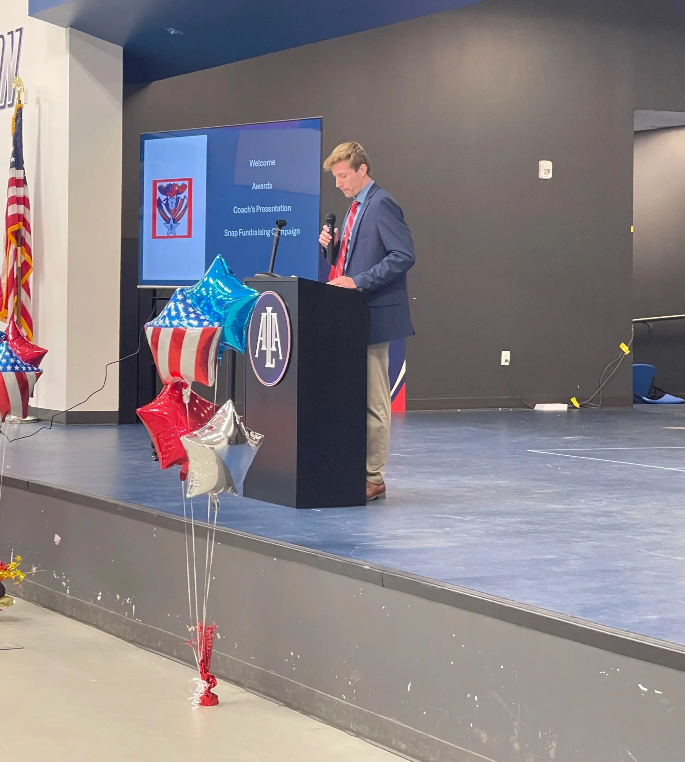A man in a suit stands at a podium decorated with an autographed logo, giving a presentation with balloons in red, white, blue, and silver with stars and stripes nearby; a large screen behind displays a welcome message and agenda for a coach's presentation and fundraising campaign.