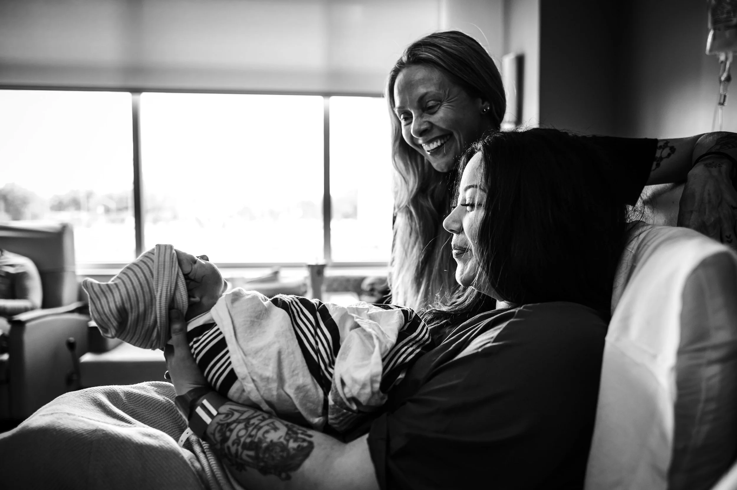 A woman holding a newborn baby in a hospital room, with a smiling woman leaning over them, all in black and white. UF North doula photography Dallas Arthur Jacksonville doulas birth