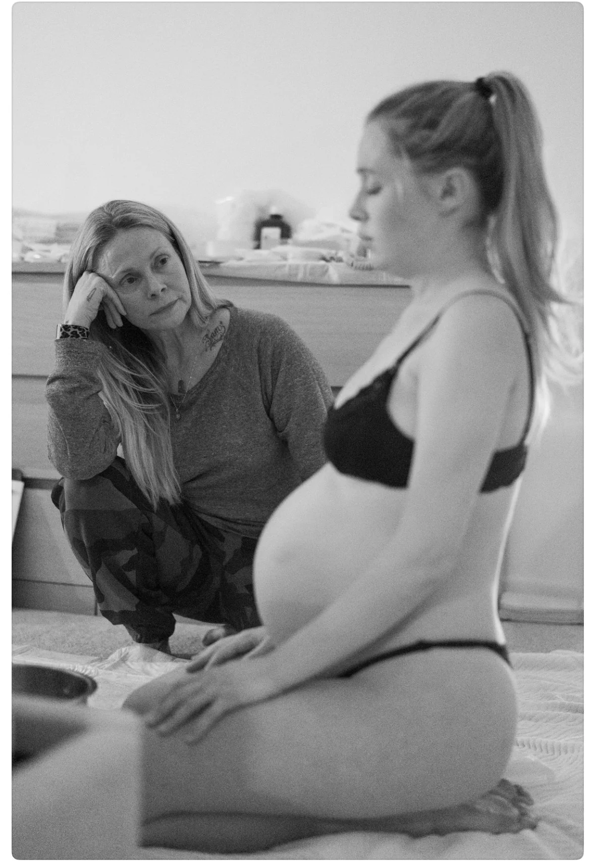 A woman sitting on the floor in front of a pregnant woman with her eyes closed and hand to her head, appearing to be in a moment of reflection during labor.