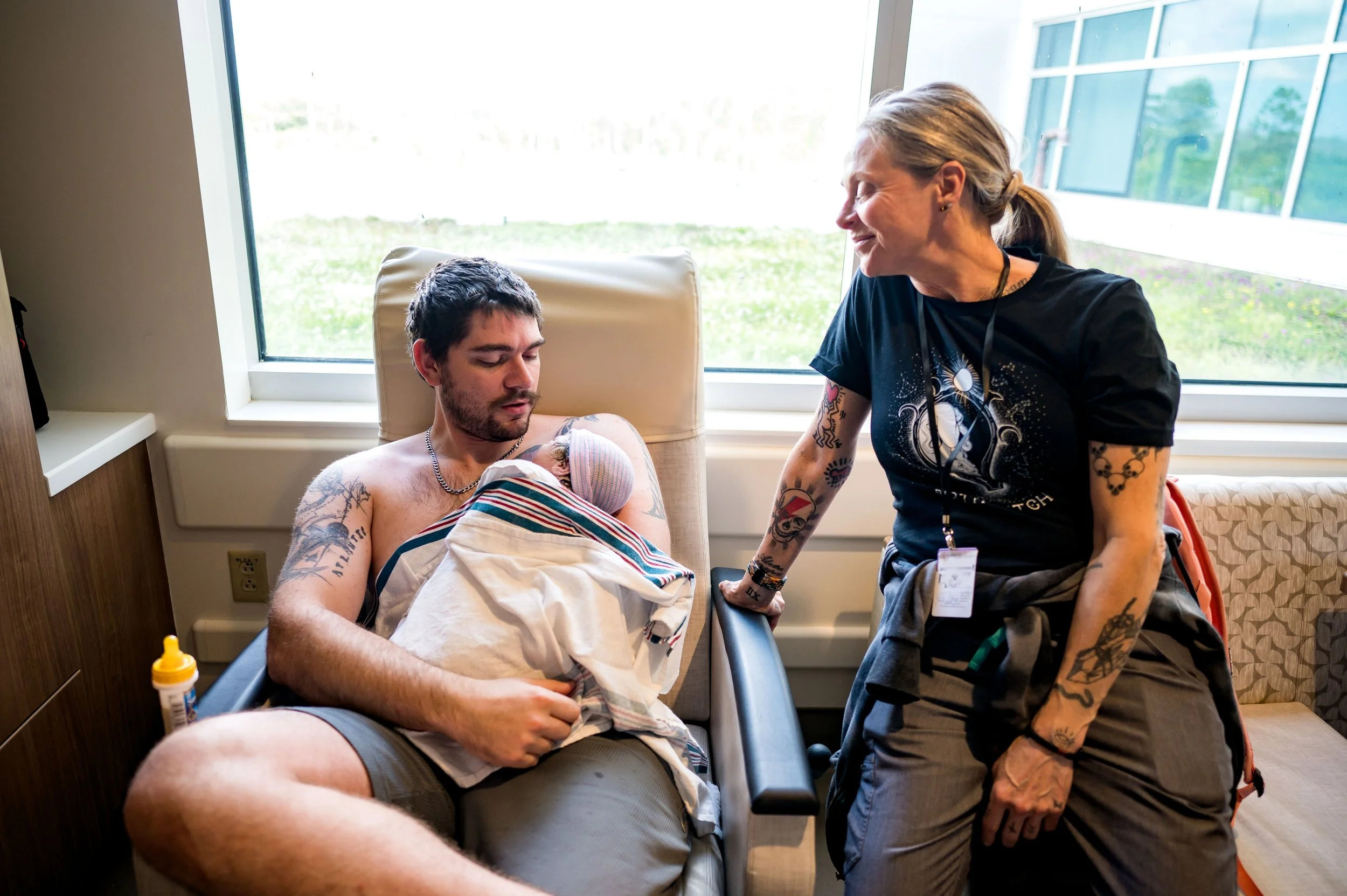 A new father holding his newborn baby in a hospital room, smiling at a nurse or hospital staff member, with bright natural light coming through the window behind them. UF North unmedicated birth 2nd time mom birth support Jacksonville Florida