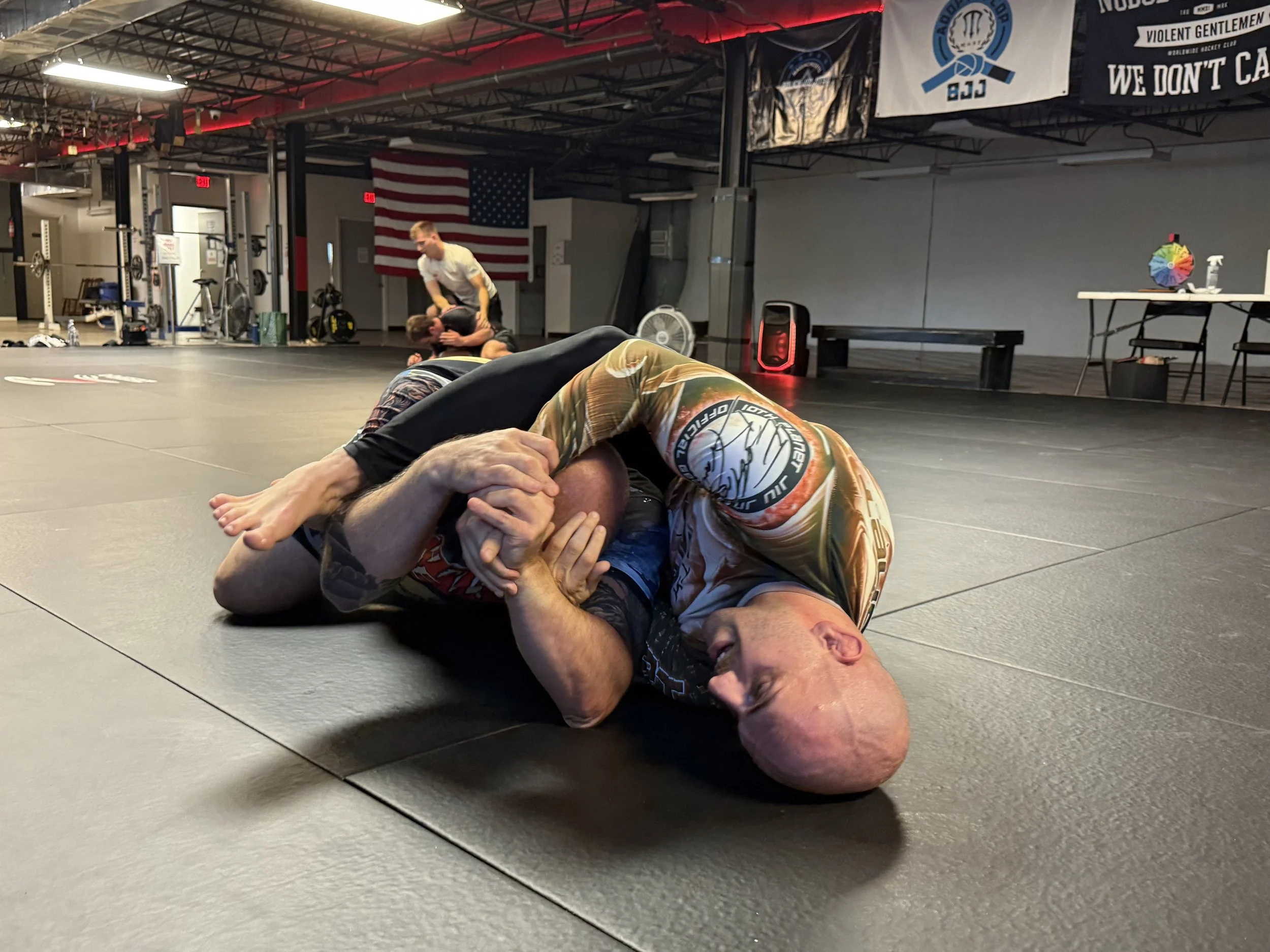 Two men practicing Brazilian Jiu-Jitsu on a mat in a gym, one in a dominant position, the other trying to defend with a submission hold. In the background, two other fighters sparring and training equipment visible.