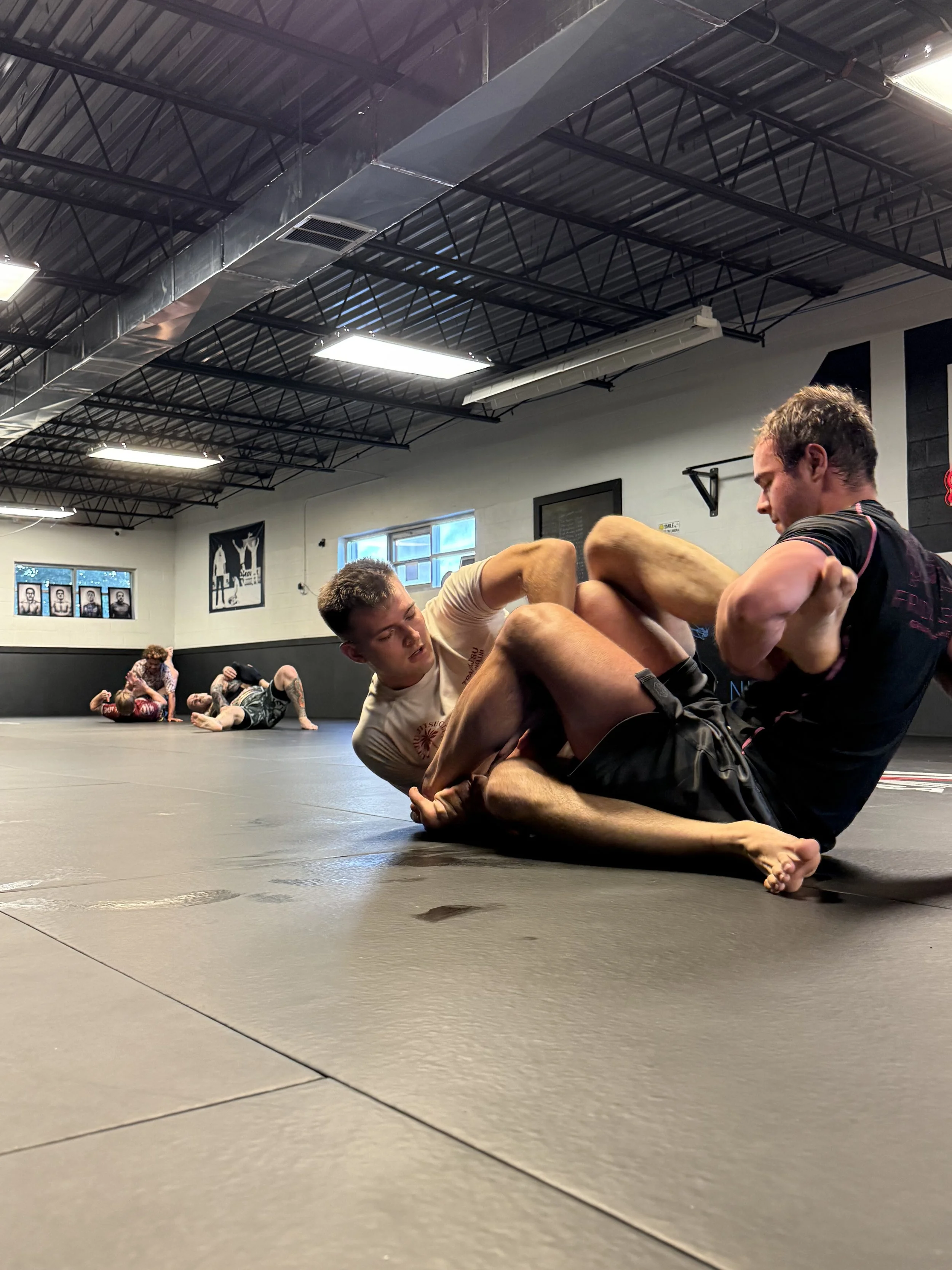 Two men practicing Brazilian Jiu-Jitsu on a gray mat in a martial arts gym, with others training in the background.