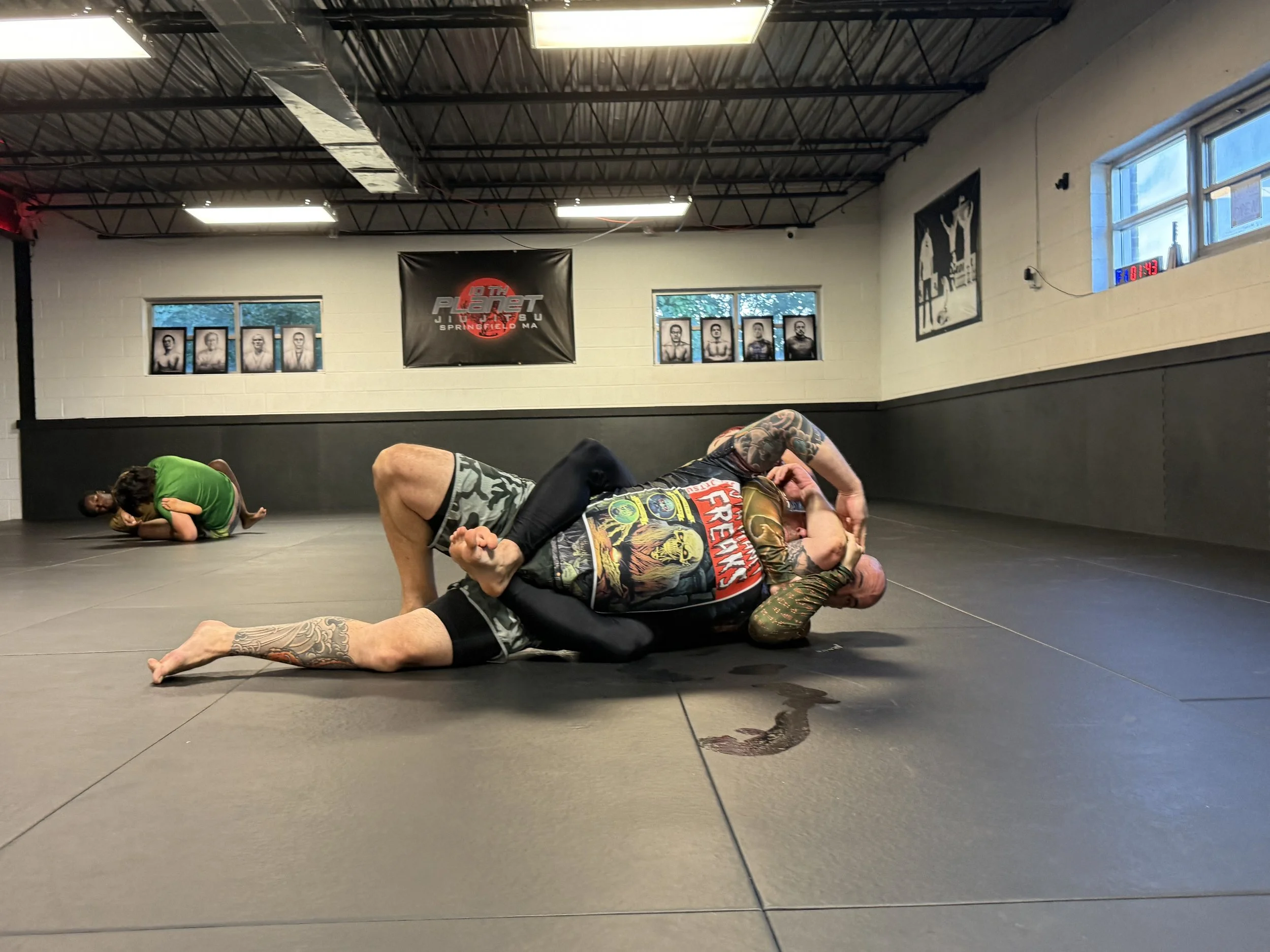 Two men practicing Brazilian Jiu-Jitsu grappling while a young boy in a green shirt watches in the background inside a martial arts gym with black padded walls and posters on the wall.