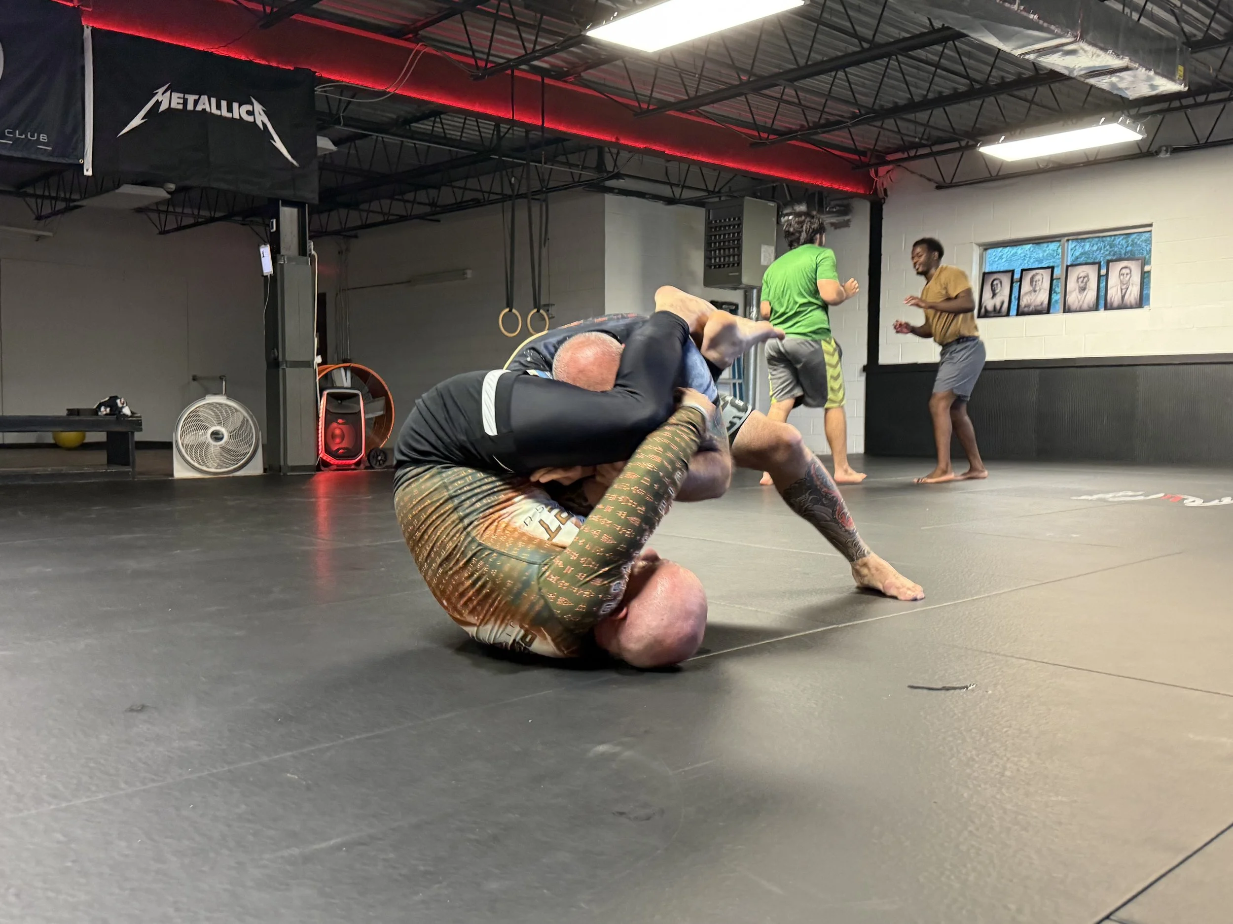 Two people grappling on the mat during Brazilian Jiu-Jitsu training, with three other individuals standing and practicing in the background inside a gym.