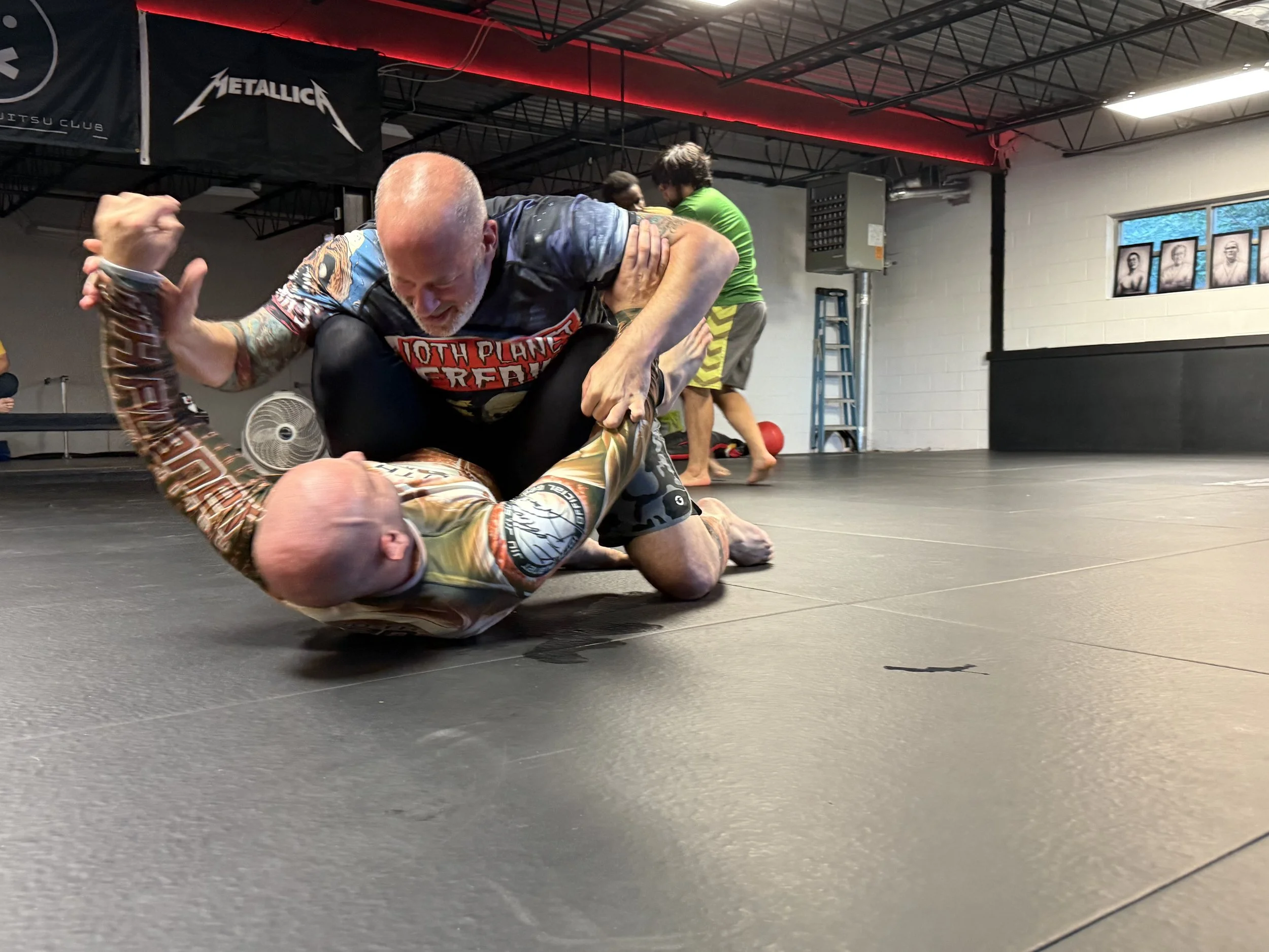Two men practicing Brazilian Jiu-Jitsu on a black mat in a gym, with one man on top and the other on the ground in a defensive position, surrounded by other practitioners and gym equipment.