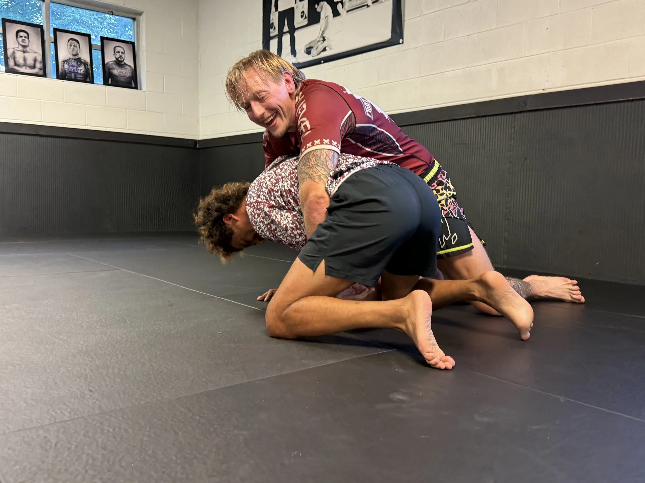 Two men practicing Brazilian Jiu-Jitsu on a black mat in a gym. One man is on top, smiling, wearing a maroon rash guard and black shorts, while the other man is on his knees and hands, wearing a patterned rash guard and black shorts. The gym walls ha