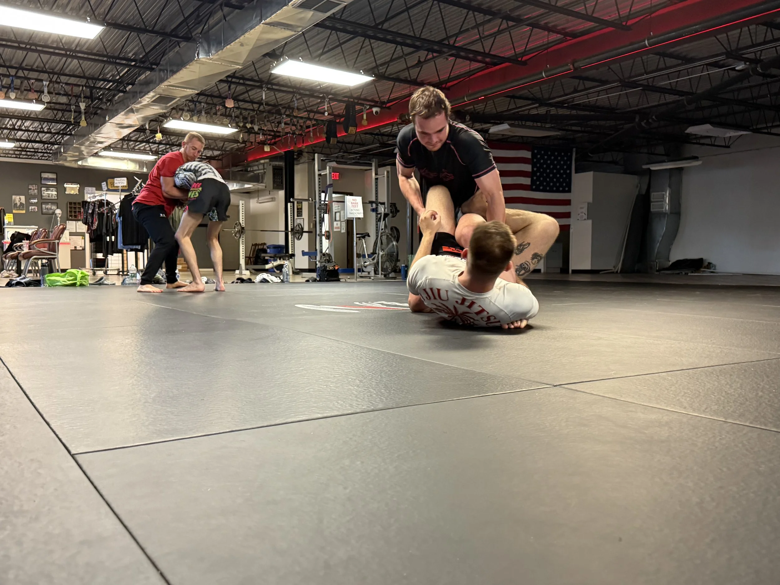 Brazilian Jiu-Jitsu practitioners training on a black mat in a gym; one person is on the ground gripping another's leg while a trainer assists nearby, with other practitioners sparring in the background.