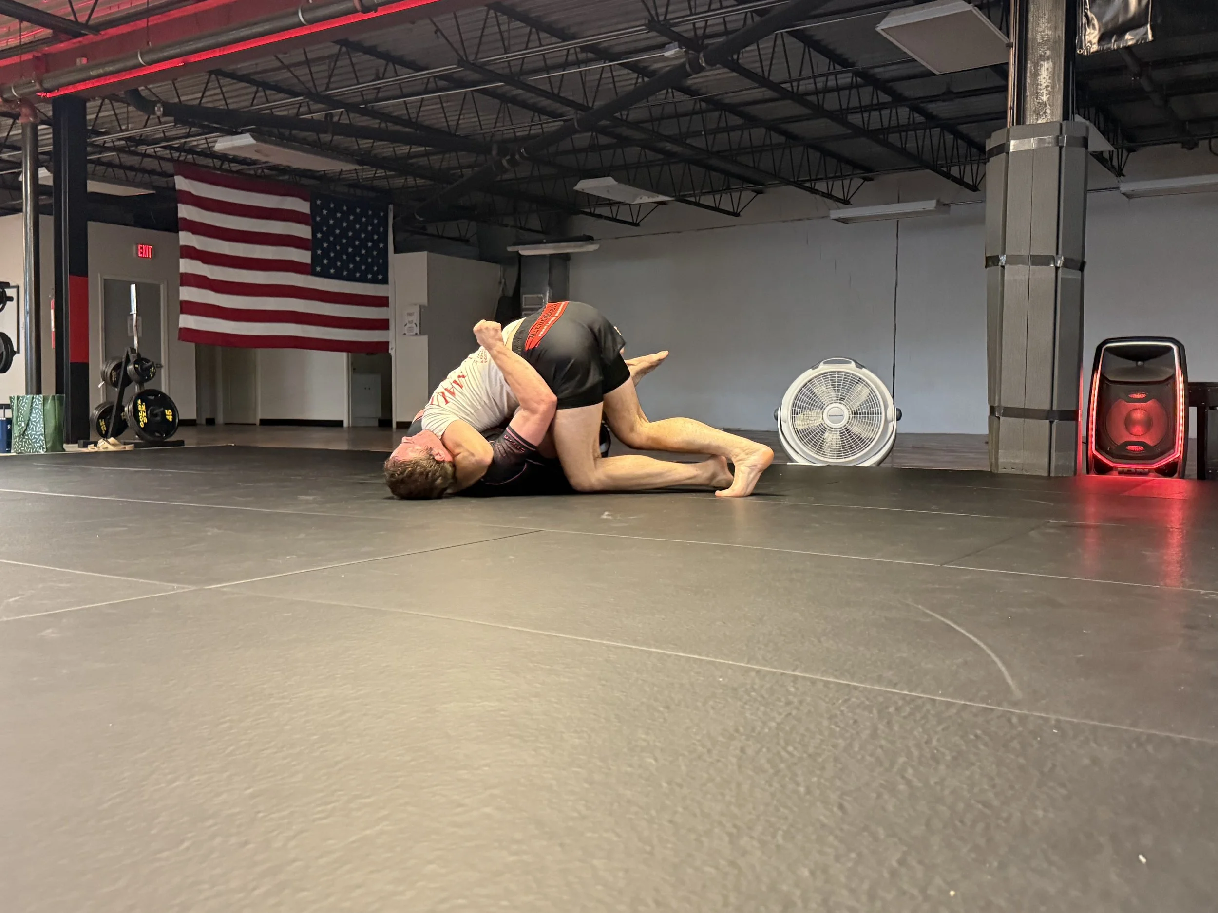 Two people practicing Brazilian Jiu-Jitsu on a mat in a gym with an American flag on the wall, weights in the background, and a large fan facing them.