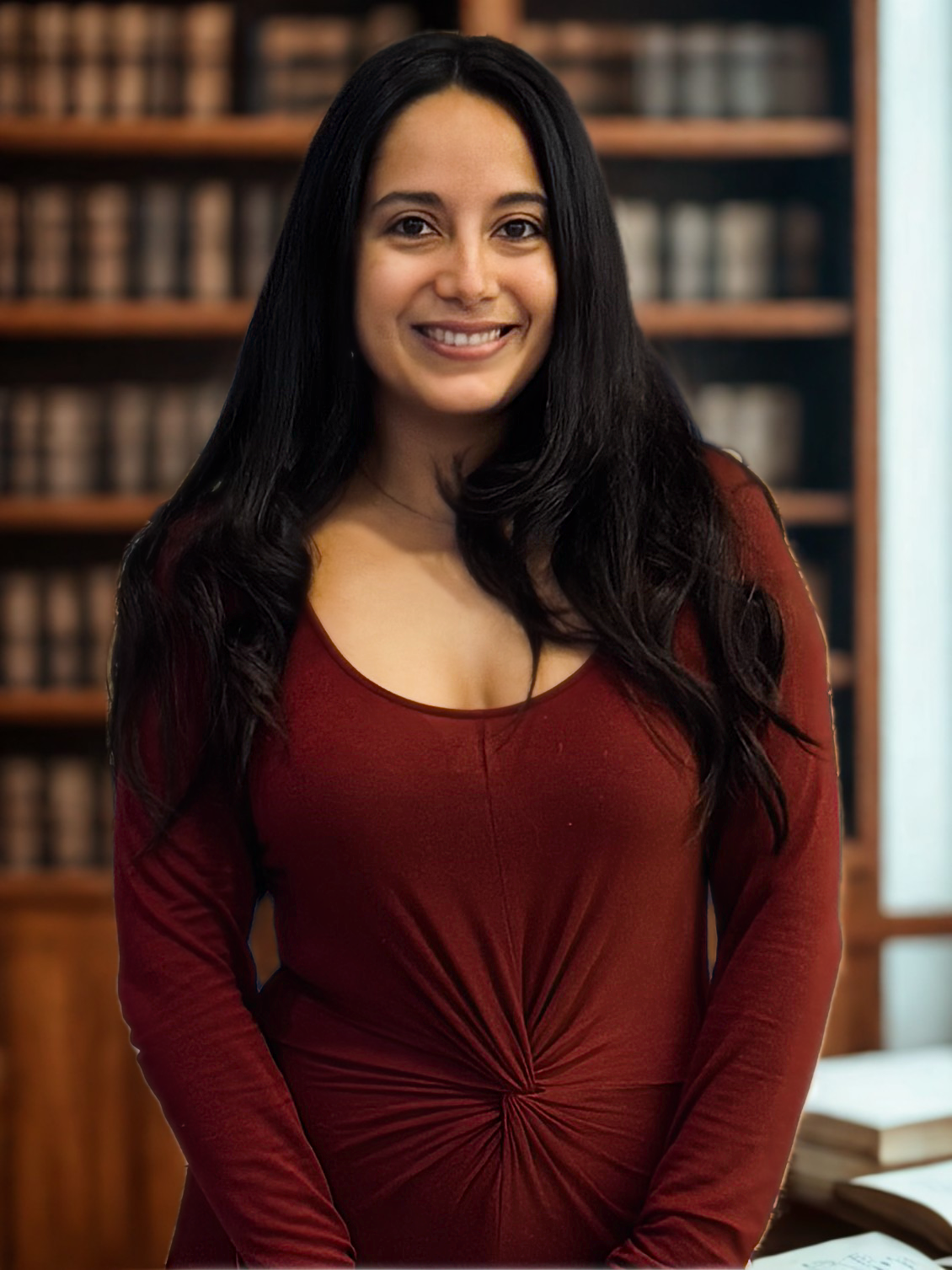 A woman with long black hair, smiling, wearing a red long-sleeve dress, stands in front of a bookshelf filled with books in a library setting.