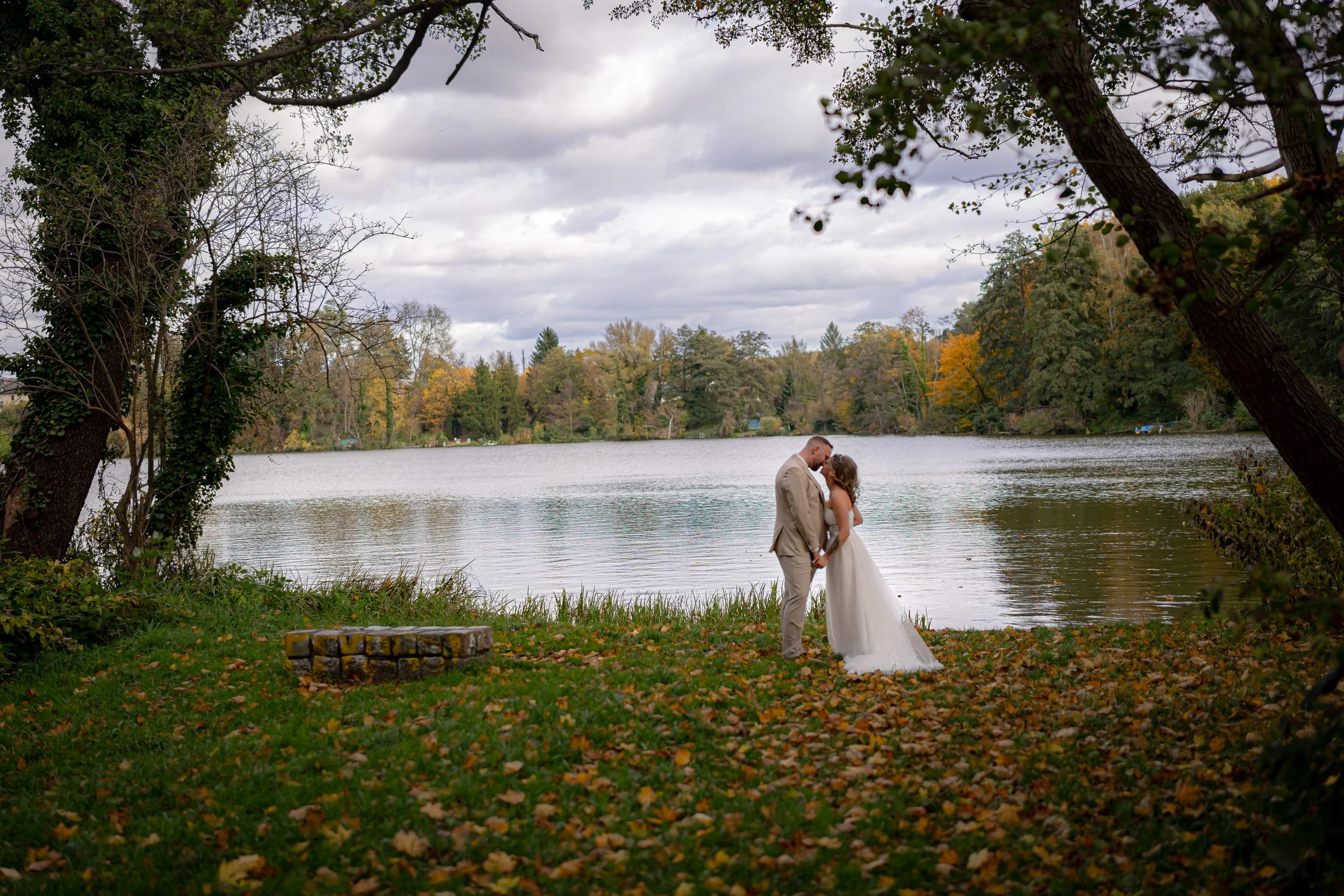Ein Brautpaar, das sich an einem See im Herbst küsst, umgeben von bunten Bäumen und fallendem Laub.