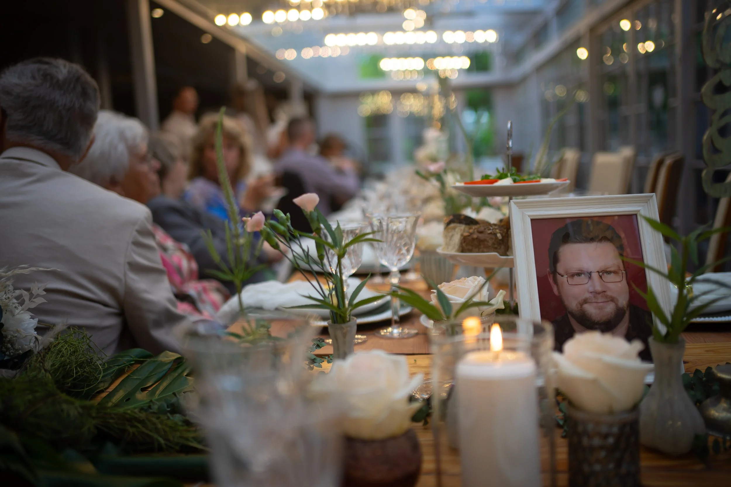 Ein gedeckter Tisch bei einer Gedenkfeier mit einem Porträt eines Mannes, Kerzen und Blumen, im Hintergrund Menschen bei einer Zusammenkunft.