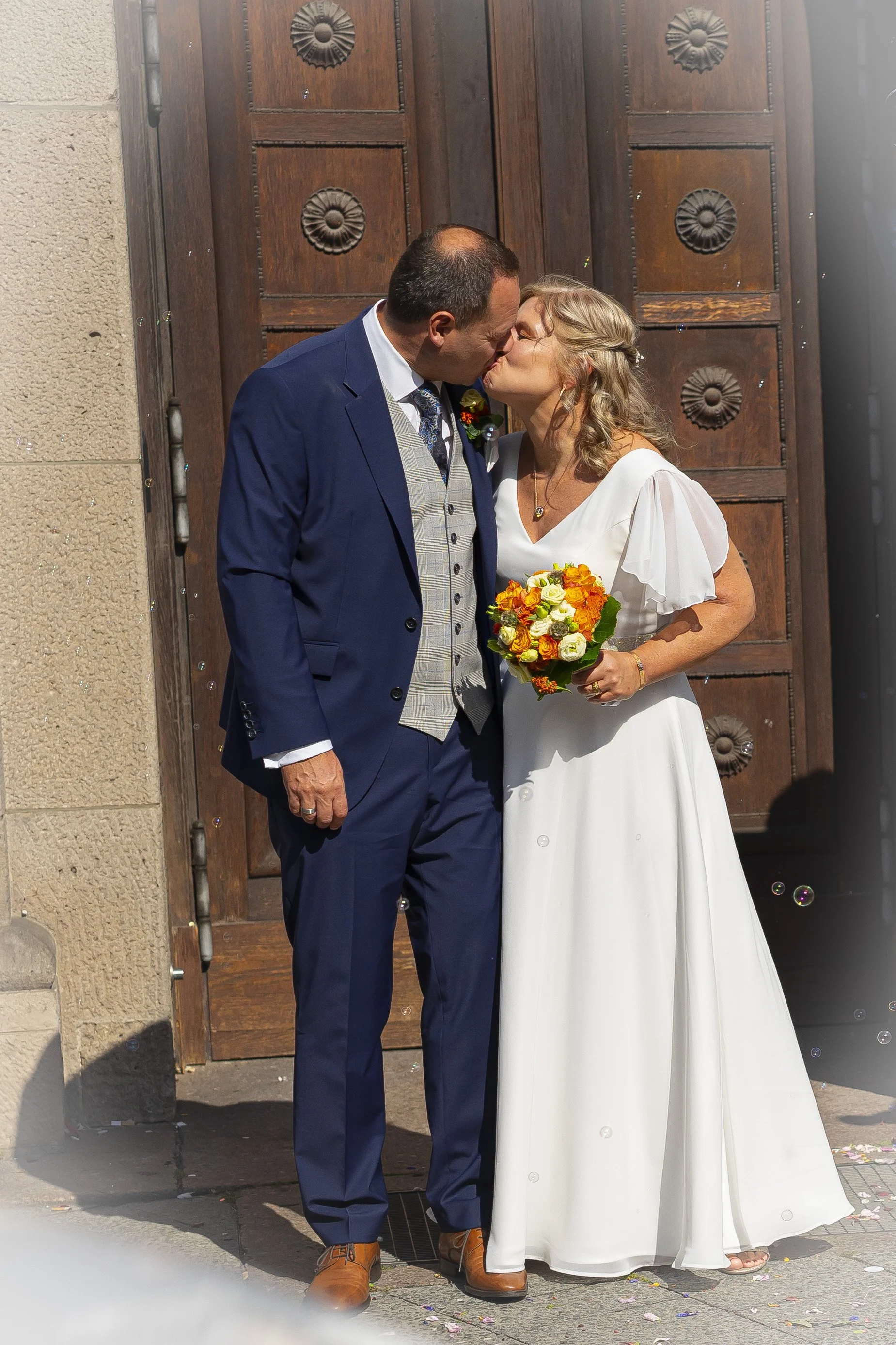 Ein Brautpaar küsst sich vor einer Holztür, die für eine Hochzeit geschmückt ist, die Braut trägt ein weißes Kleid und hält einen Blumenstrauß, der Bräutigam trägt einen dunklen Anzug.