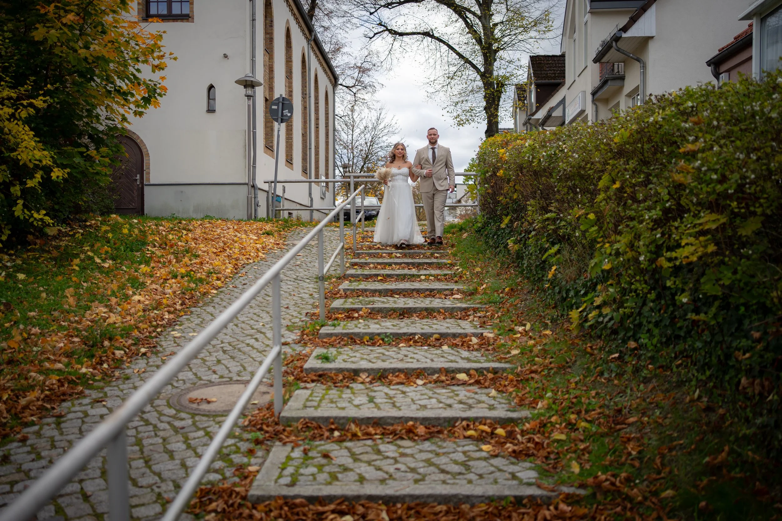 Ein Brautpaar in Hochzeitskleidung läuft die Treppe eines Gehwegs in einer Stadt hinauf, umgeben von Herbstlaub, Büschen und Gebäuden.