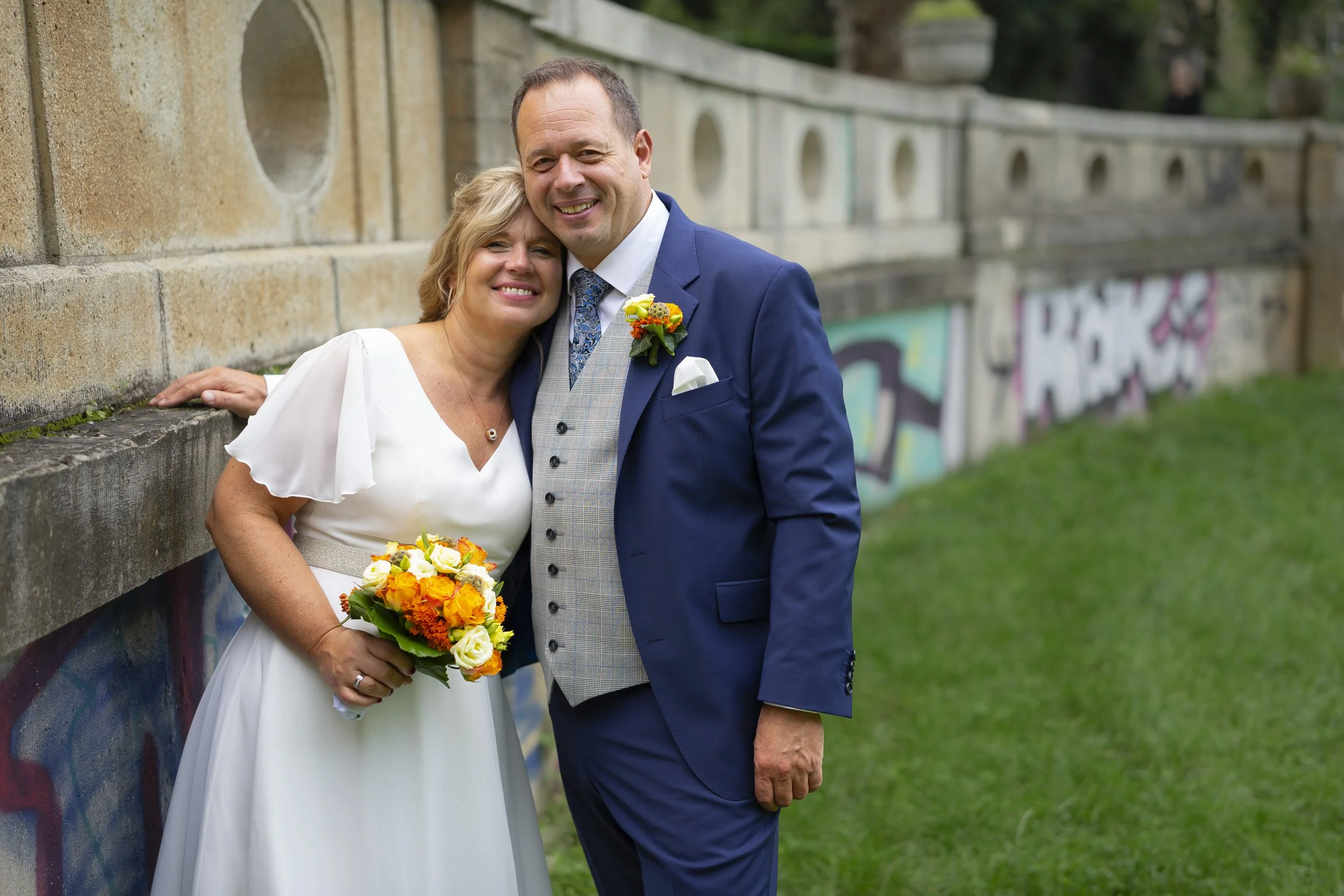 Ein Paar bei einer Hochzeit, die Frau hält einen Blumenstrauß, beide lachen und stehen eng zusammen vor einer Graffiti-Wand im Freien.