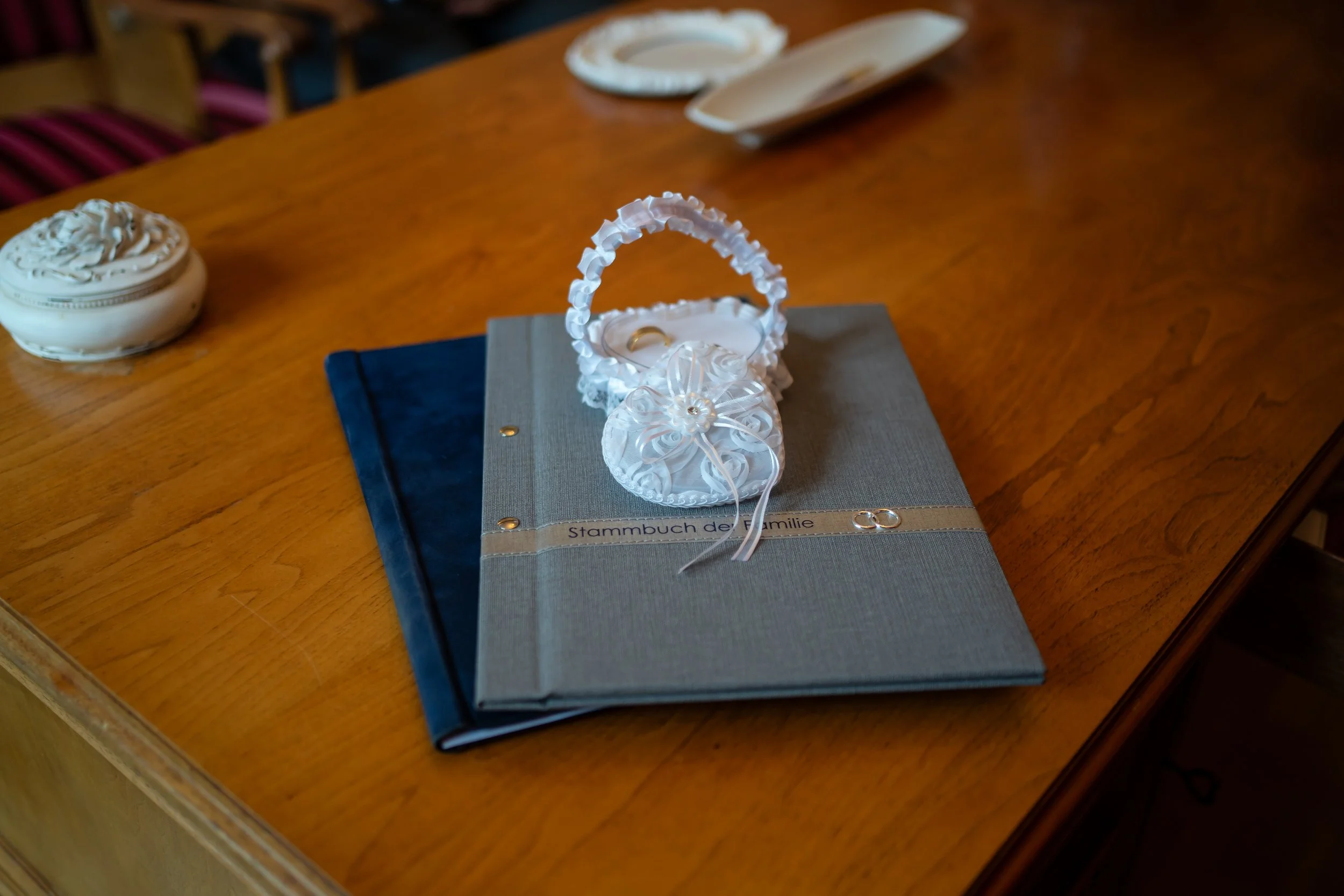 Ein Archivordner mit dem Titel "Stammbuch der Familie" liegt auf einem Holz Tisch, darüber liegt eine kleine, weiße, verzierte Babytasche mit einer Schleife und Perlen. Im Hintergrund sind Papierteller und eine Schale zu sehen.