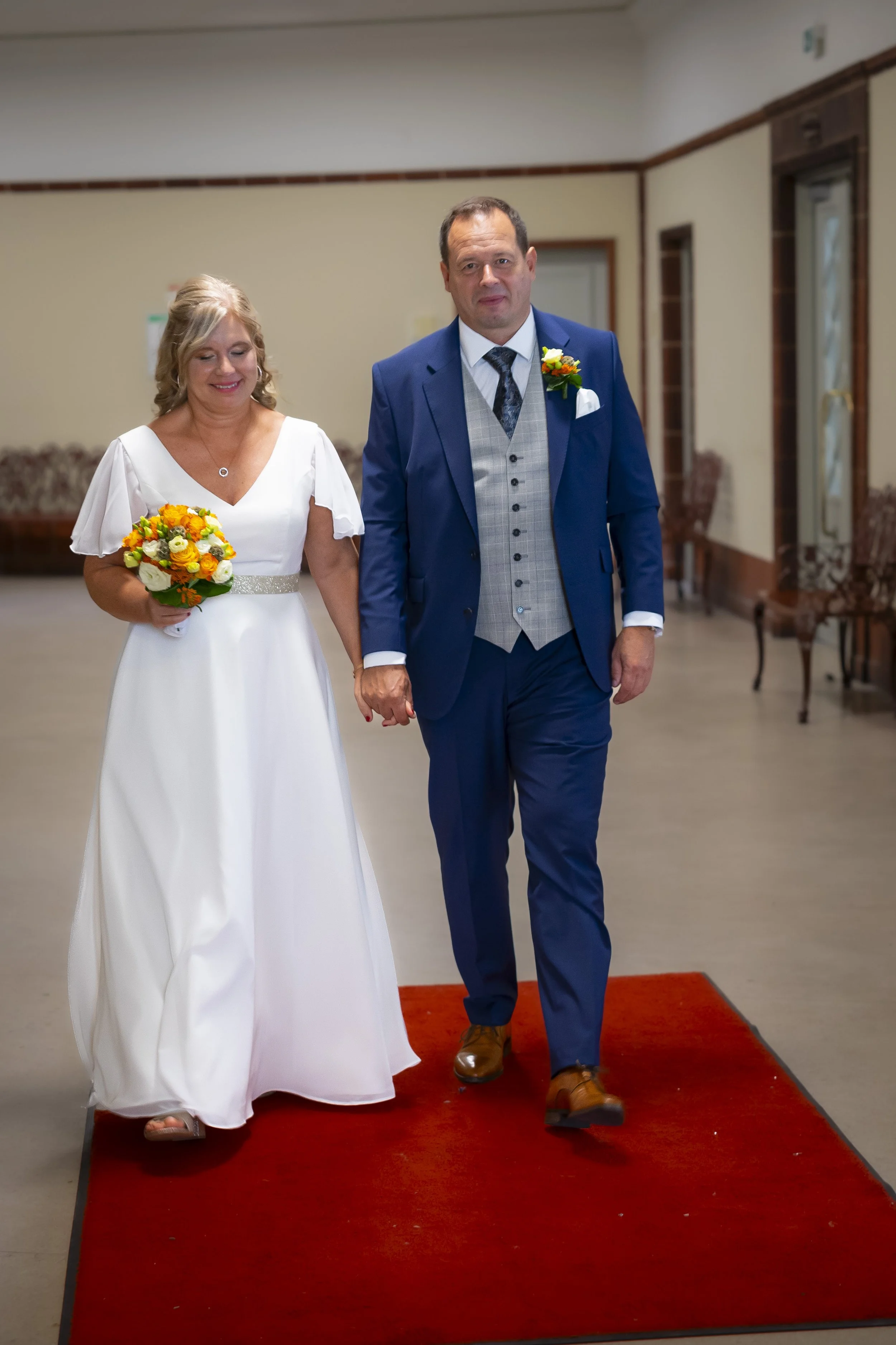 Ein Brautpaar geht Hand in Hand auf einem roten Teppich während einer Hochzeitszeremonie. Die Braut trägt ein weißes Kleid und hält einen bunten Blumenstrauß, der Bräutigam trägt einen blauen Anzug.