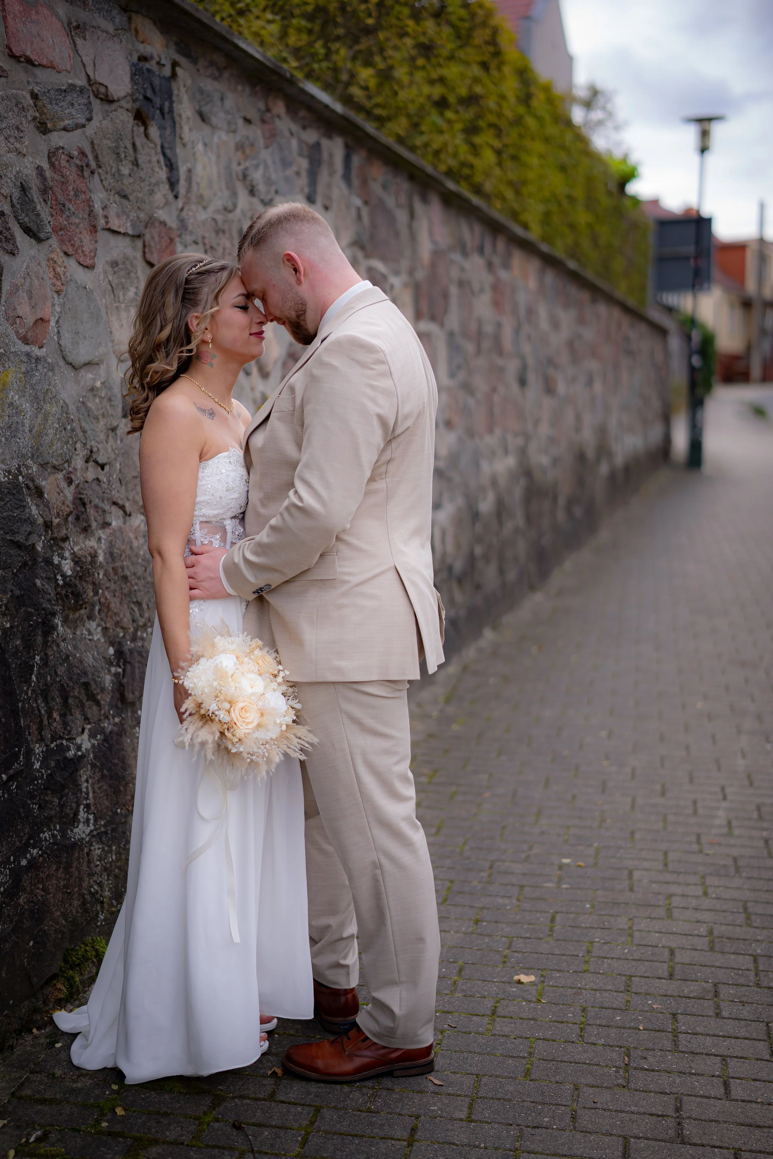 Ein Brautpaar steht an einer Steinmauer auf einem Bürgersteig, sie berühren sich mit der Stirn und schauen sich liebevoll an, es ist ein regnerischer Tag.