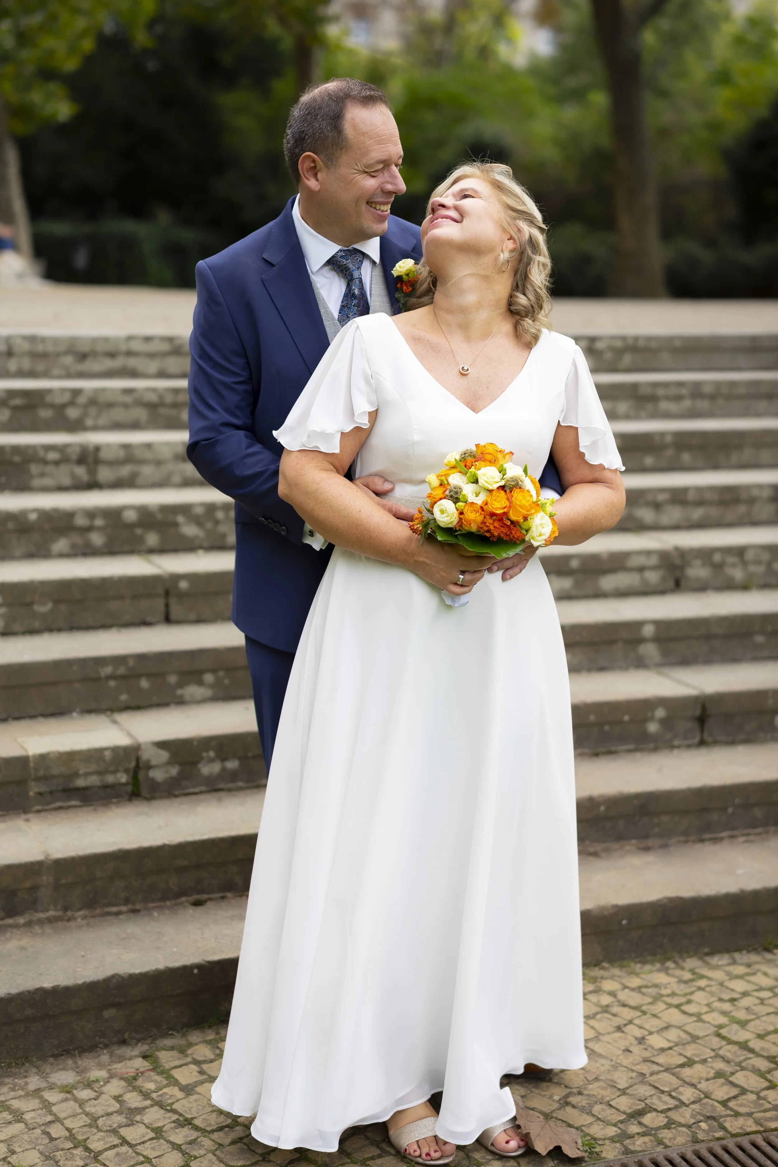 Ein glückliches Brautpaar bei einer Hochzeit im Freien, der Mann trägt einen dunklen Anzug, die Frau ein weißes Kleid und hält einen bunten Blumenstrauß.