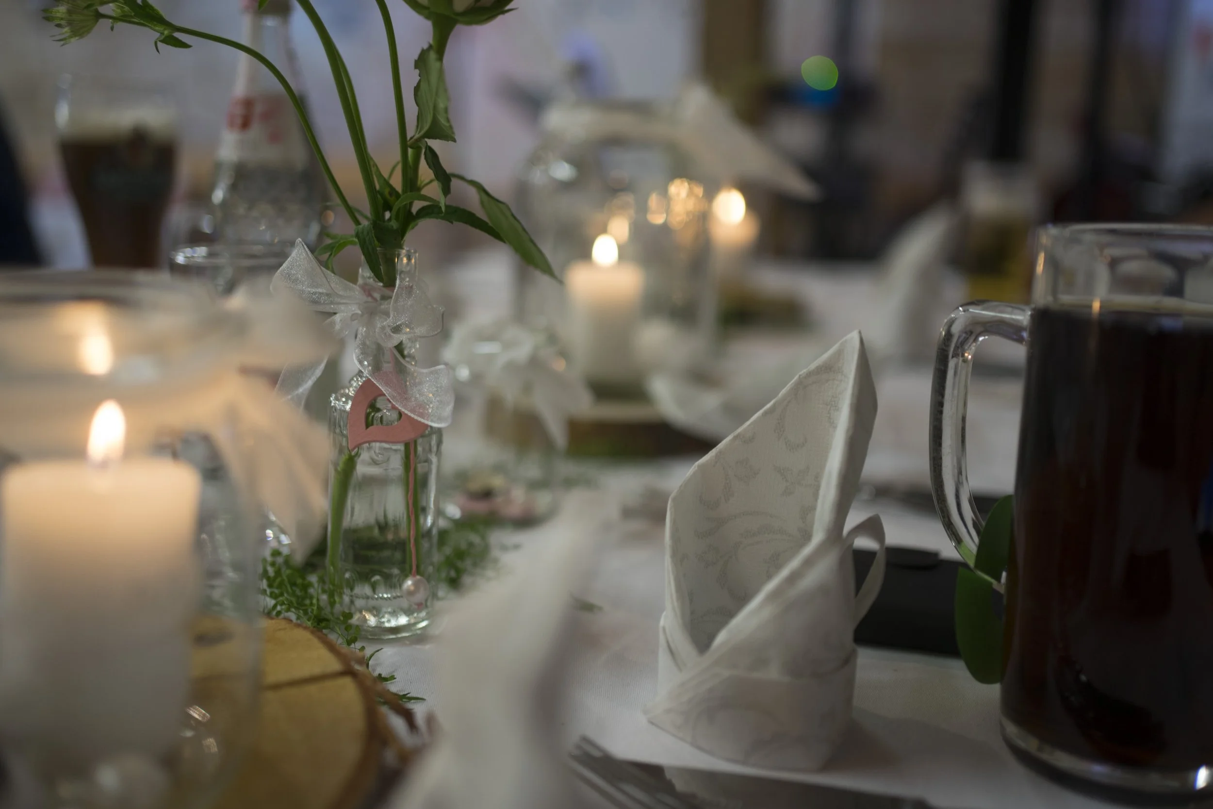 Gesicherter festlicher Esstisch mit Kerzen, weißen Stoffservietten, einer weißen Baum- oder Blumen-Dekoration in einem Glas, und einem Glas Bier sowie einem Krug mit dunklem Getränk.