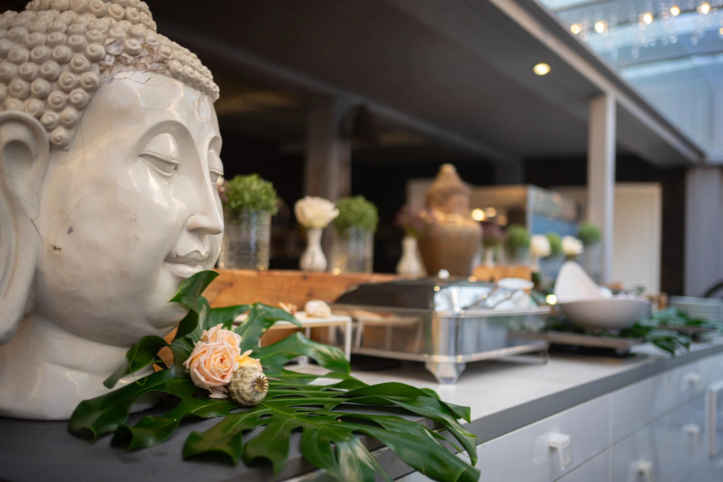 Nahaufnahme eines weißen Buddha-Kopf-Skulptur mit grünen Blättern und Blumen im Vordergrund, im Hintergrund Küchenutensilien und Dekorationen.