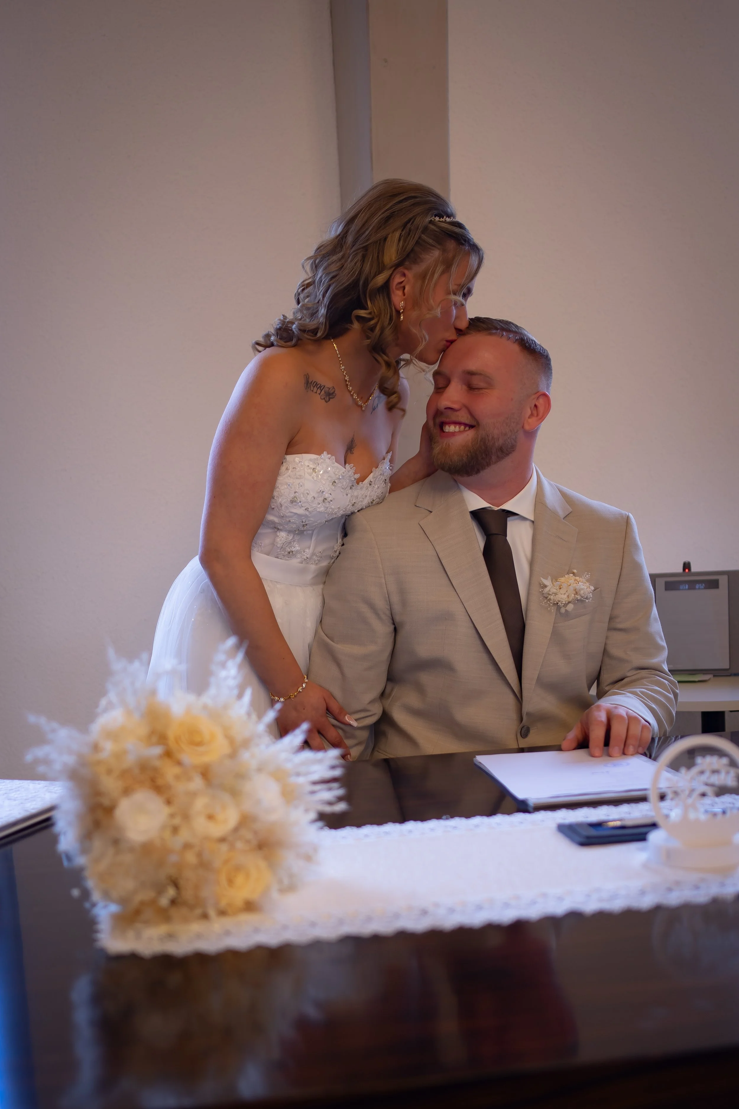 Ein Brautpaar bei der Hochzeitszeremonie. Die Braut küsst den Bräutigam auf die Stirn, während er lächelnd auf dem Stuhl sitzt. Im Vordergrund liegt ein Brautstrauß auf einem Tisch.