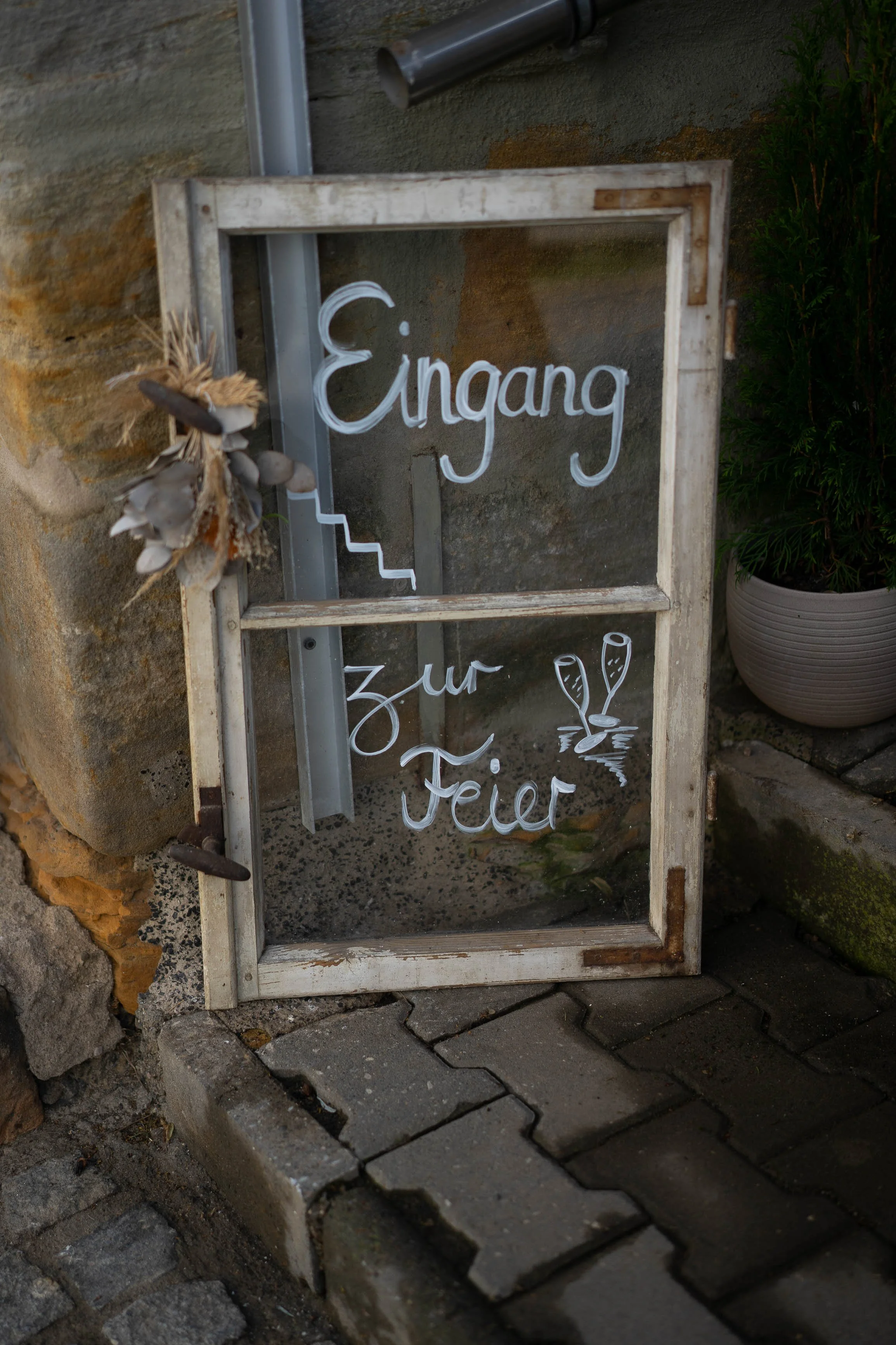 Ein handgeschriebenes Schild mit der Aufschrift 'Eingang zur Feier' in German, dekoriert mit Deko-Elementen und an einem Steinmauer neben einem Pflanzkübel aufgestellt.